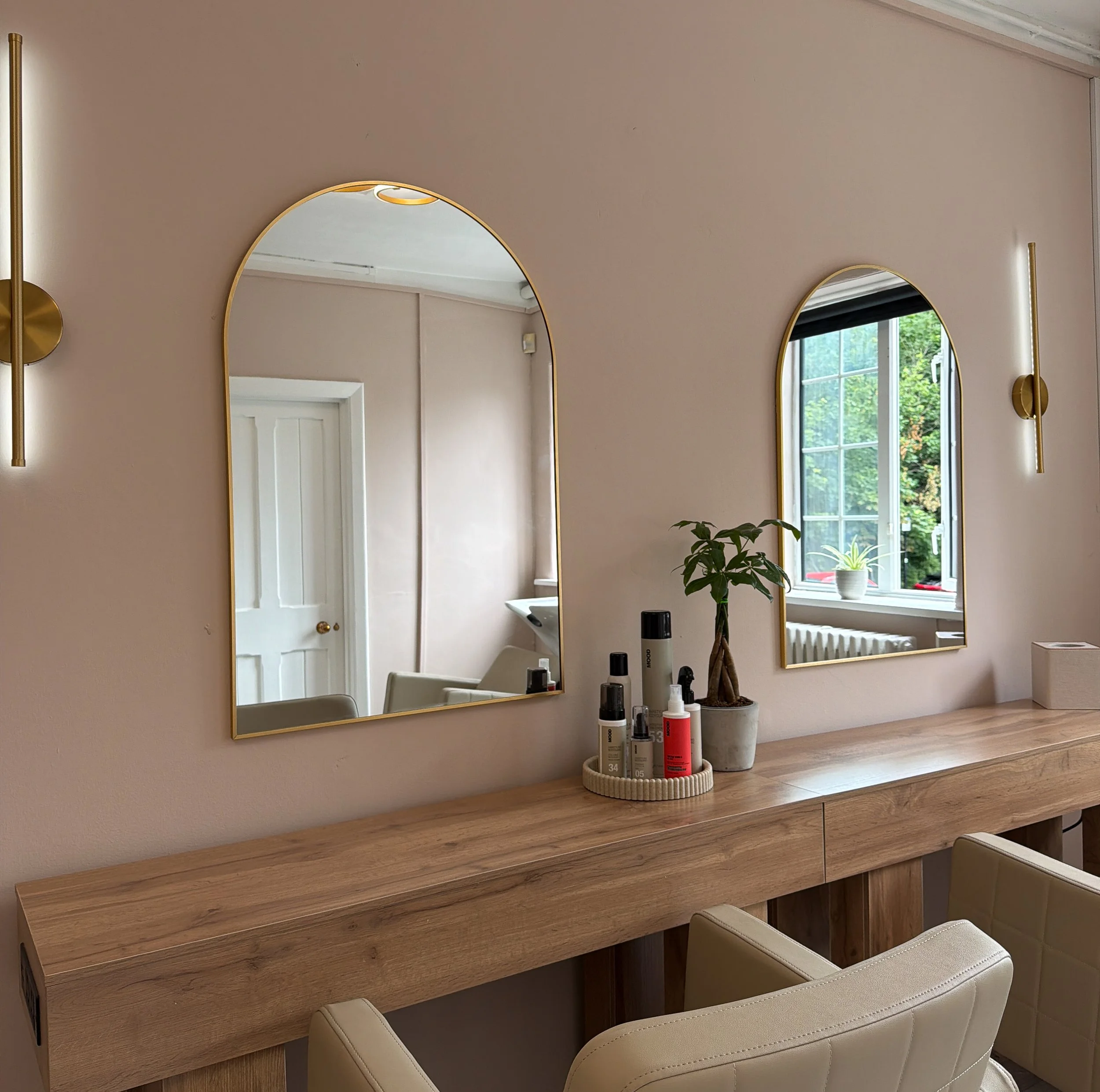 Interior view of a room with two mirrors on a beige wall, a wooden desk with skincare products, a potted plant, a window with green outdoor foliage, and beige chairs.