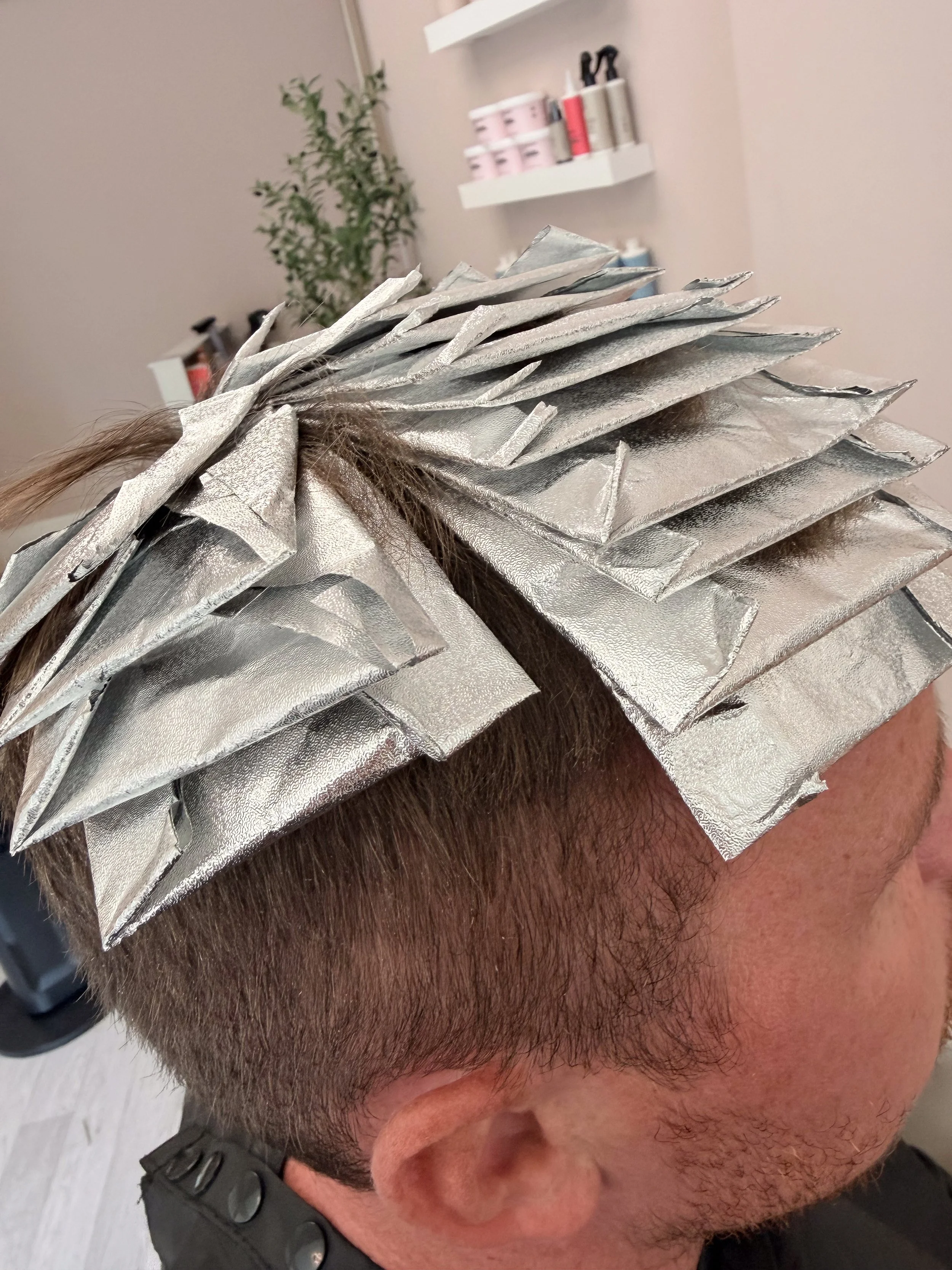 Close-up of a person's head with hair sectioned and wrapped in aluminum foil, likely for hair coloring or highlighting, with a background of salon supplies on shelves.