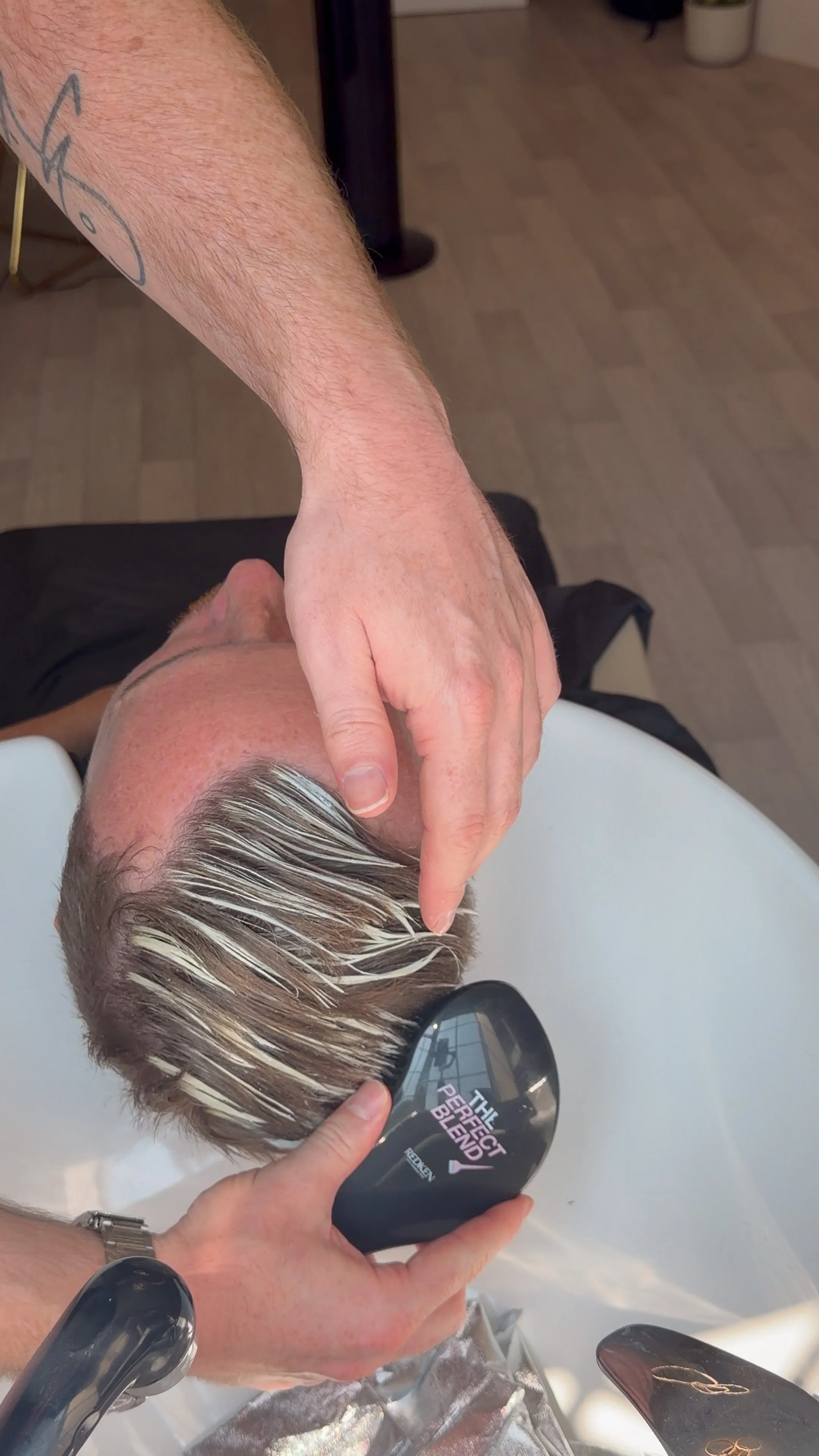 Person receiving a haircut with blonde highlighted hair, lying back in a salon chair, and a stylist's hand holding a mirror with the words 'The Perfect Blend' on it.