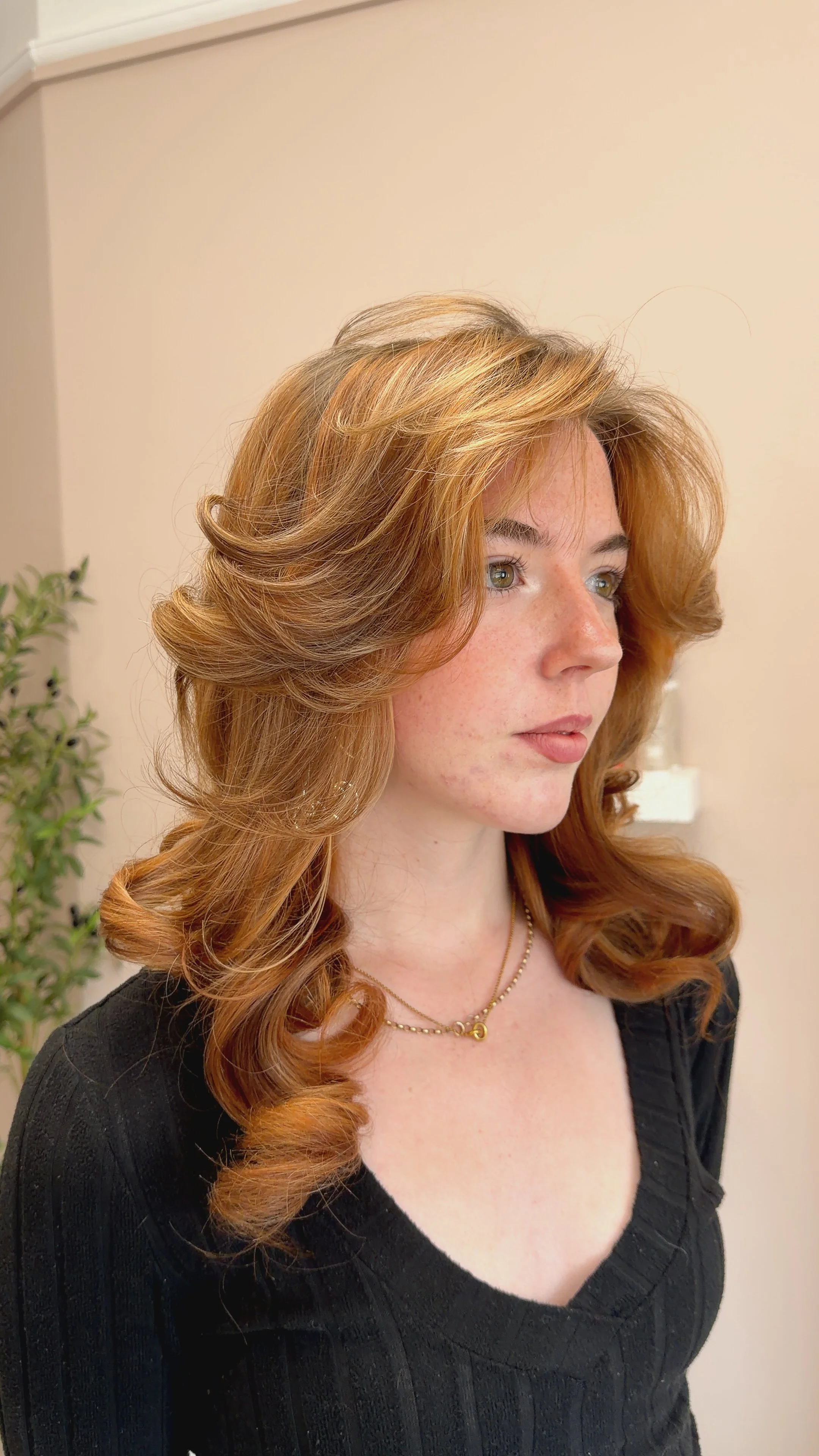 A woman with long, curly, reddish hair and light skin looks to her right. She is wearing a black top with a V-neck and a gold necklace. The background is beige with a small green plant on the left.