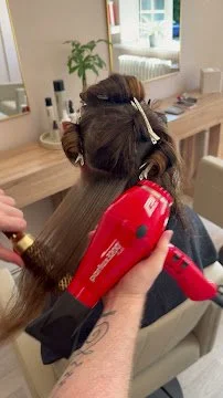 Person using a red hairdryer to dry another person's hair in a salon or home setting.