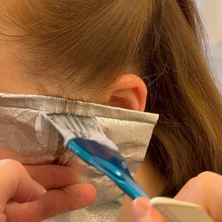 A person applies hair dye with a brush to their hair, which is partially covered with a foil cap.
