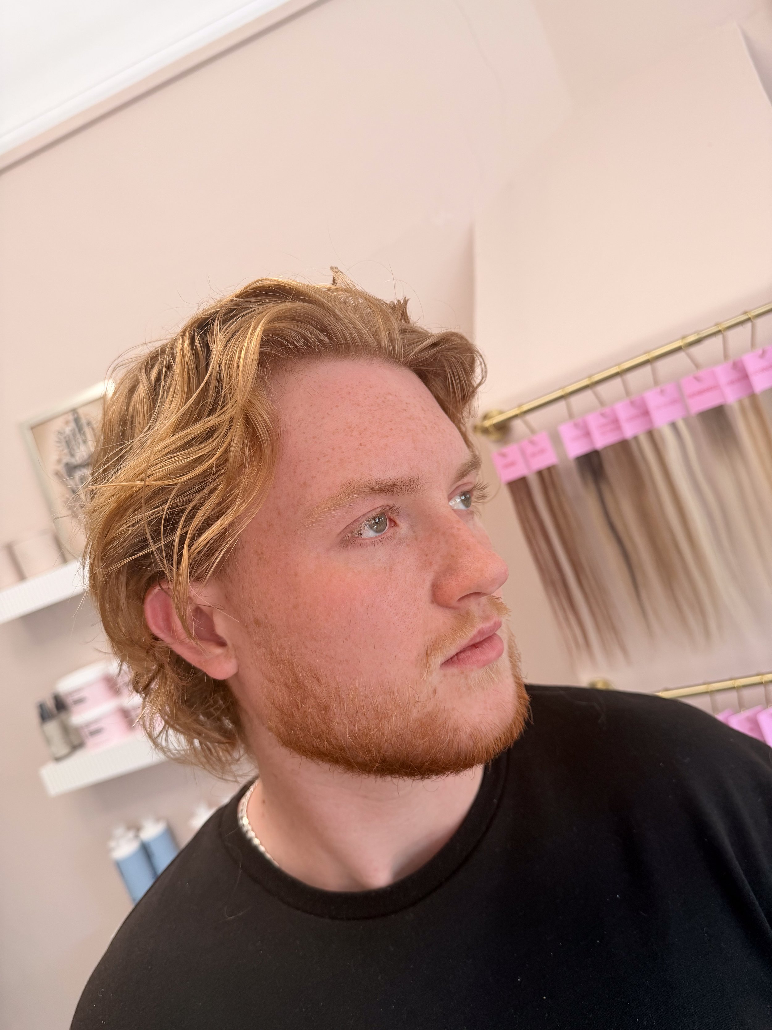 A young man with red hair, freckles, and blue eyes, wearing a black shirt and a pearl necklace, is looking away in an indoor setting with shelves and pink ribbons in the background.