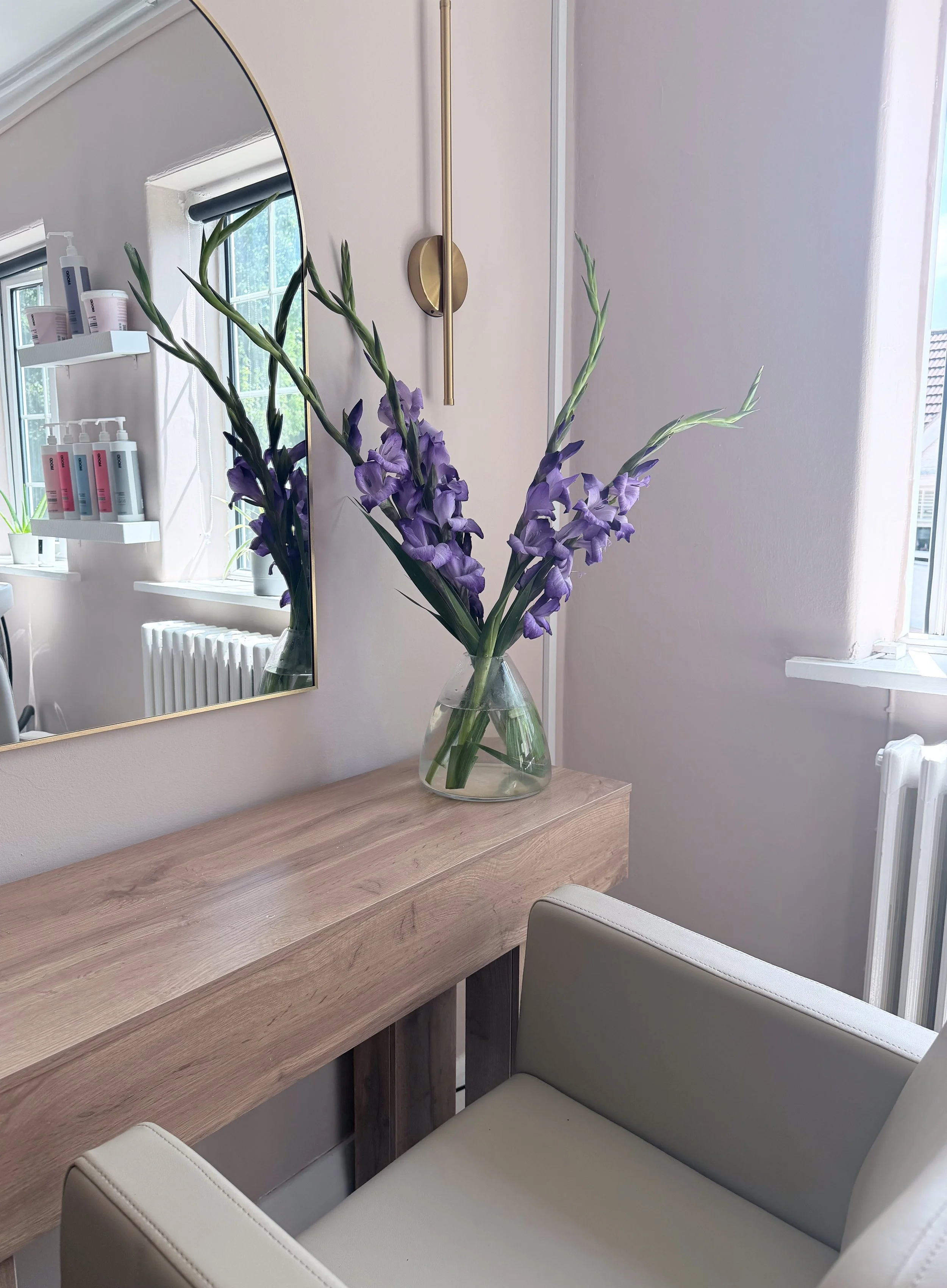 A clear glass vase with purple gladiolus flowers on a wooden desk. A white armchair with a gray backrest is partially visible in front of the desk. There is a mirror on the wall reflecting a window with trees outside and a shelf with skincare products. A metallic wall lamp is mounted above the mirror, and a radiator is below the window.