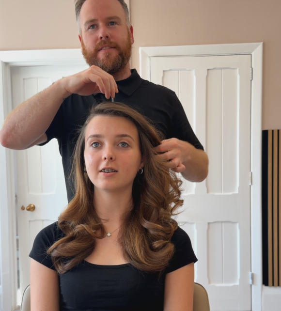 A man with a beard styling a woman’s long, wavy brown hair in a room with white doors.
