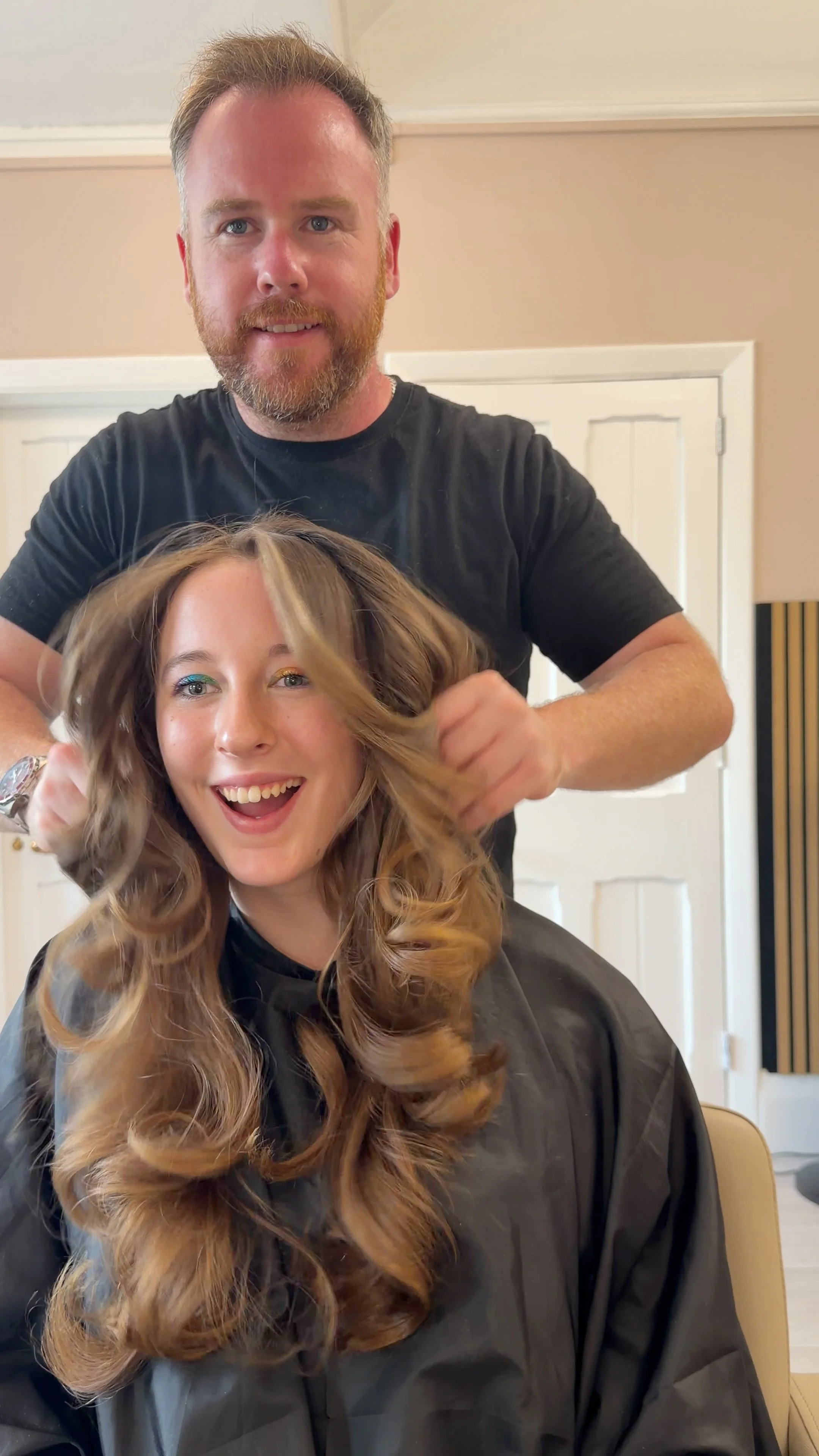A man with a beard and a woman with long curly hair are smiling inside a hair salon. The man is standing behind the woman, holding her hair.