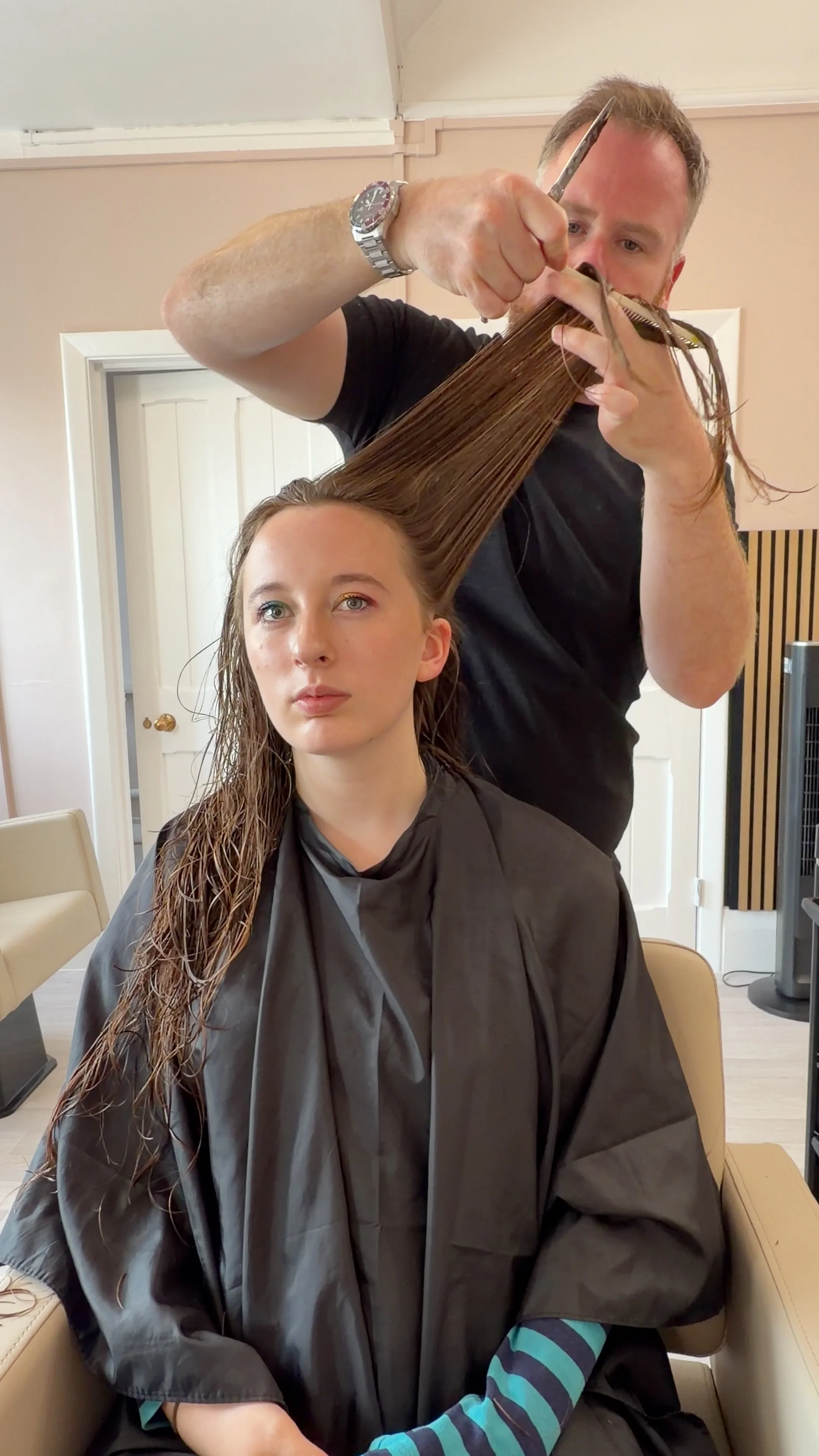 A man is cutting a young woman's hair in a salon, and she is sitting in a chair wearing a hairdressing cape. The man is holding scissors and combs, and is focused on his work.