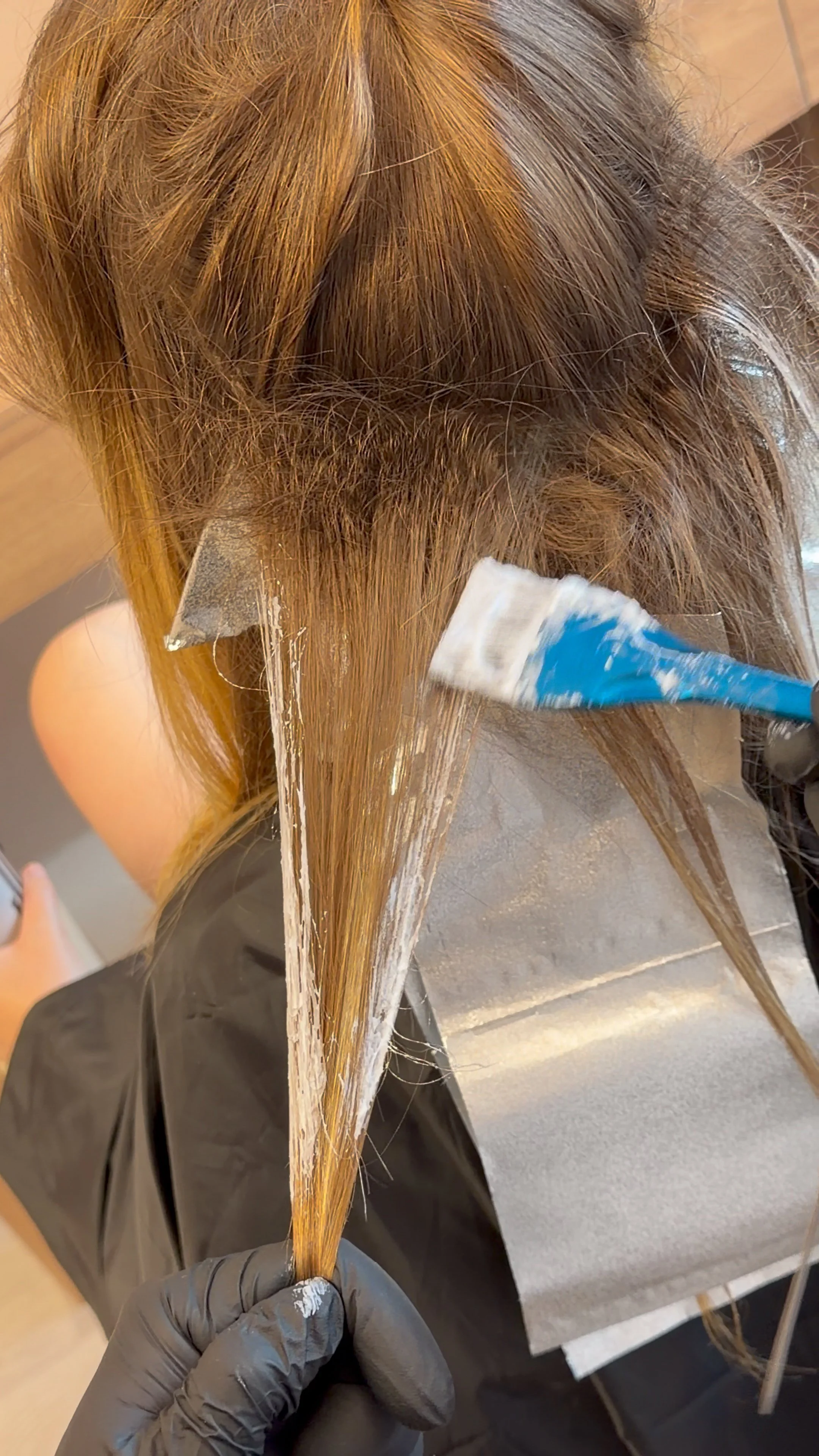 A person with long, light brown hair getting hair dye applied by a stylist wearing black gloves in a salon.