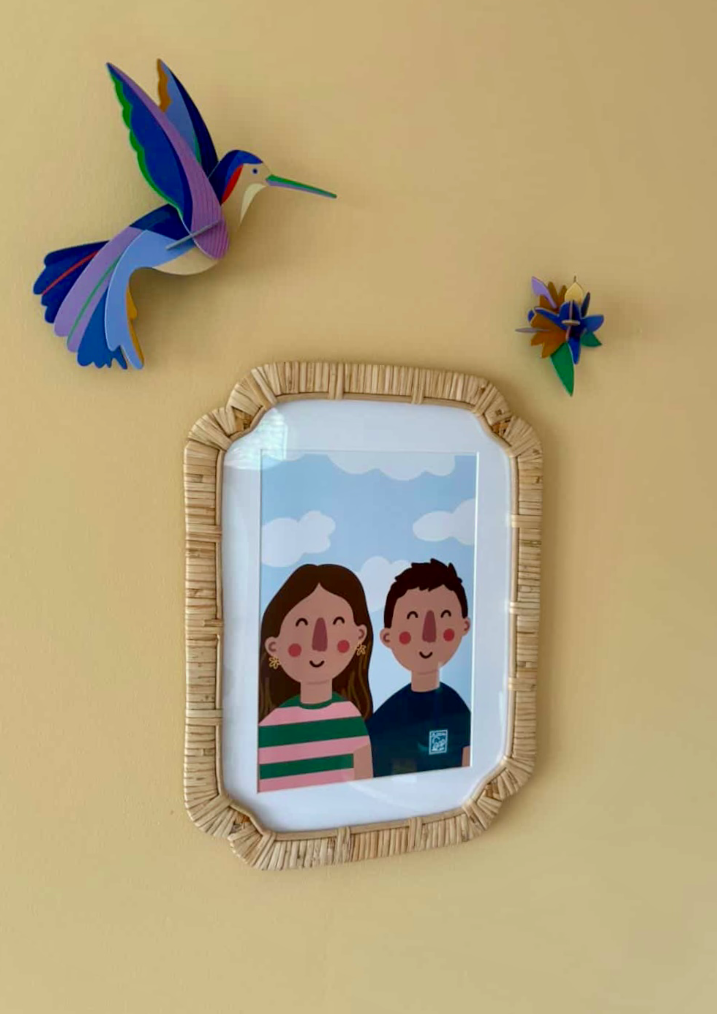 A colorful paper craft of a hummingbird and a small flower on a cream-colored wall, with a mirror with a rattan frame below.