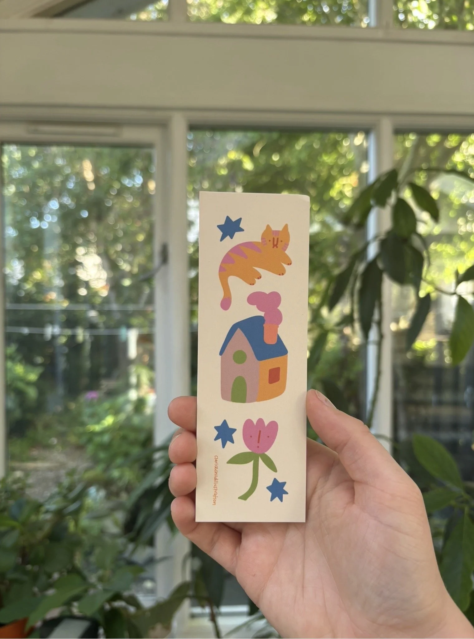 Hand holding a colorful illustrated bookmark with a house, a flower, a cat, and stars in front of a window with greenery outside.