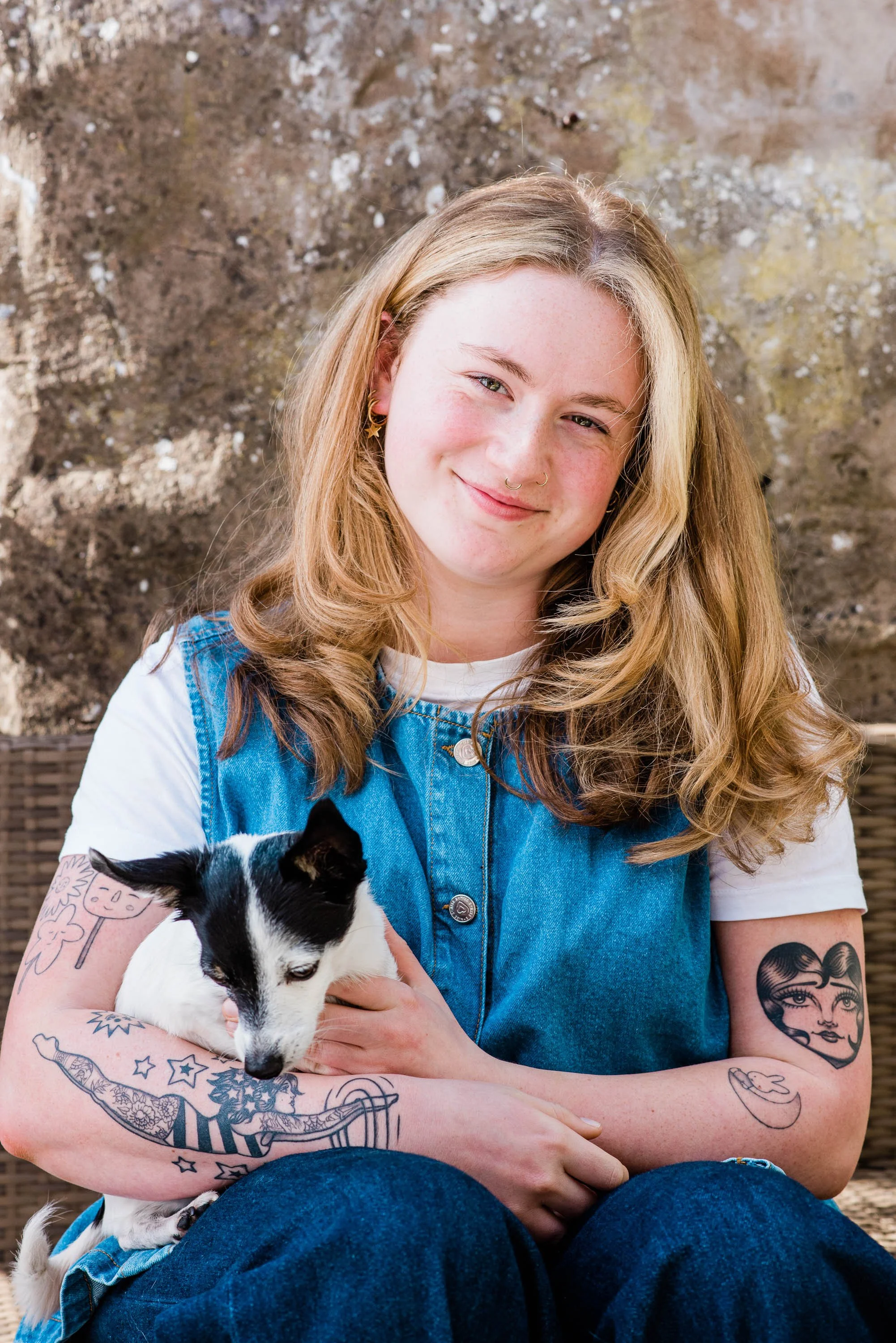 A young woman with long, wavy blonde hair and tattoos on her arms is sitting outdoors, holding a black and white puppy. She is smiling and wearing a denim vest over a white t-shirt.