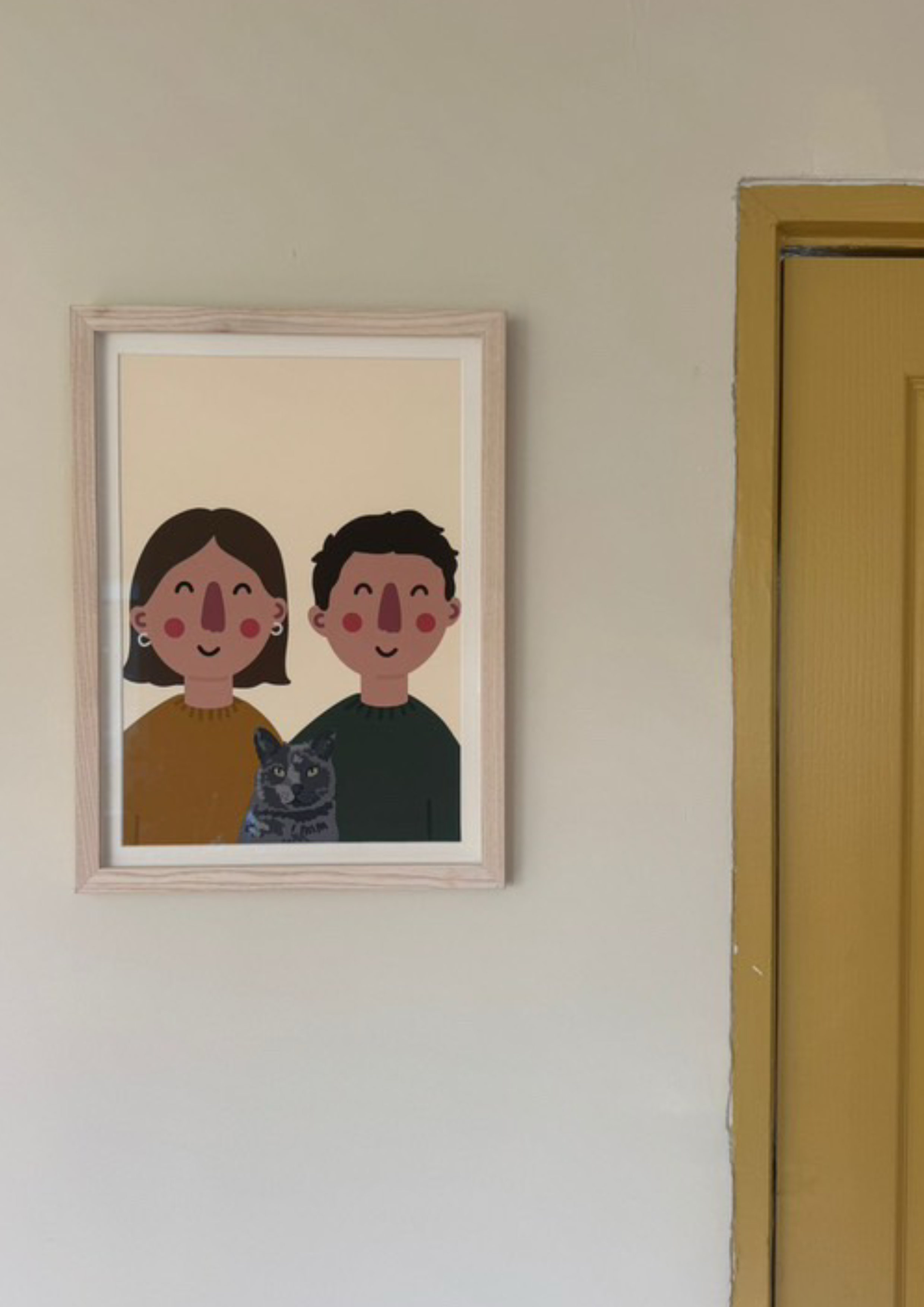 Framed illustration of a smiling woman and man with a gray cat, hung on a beige wall near a yellow doorframe.