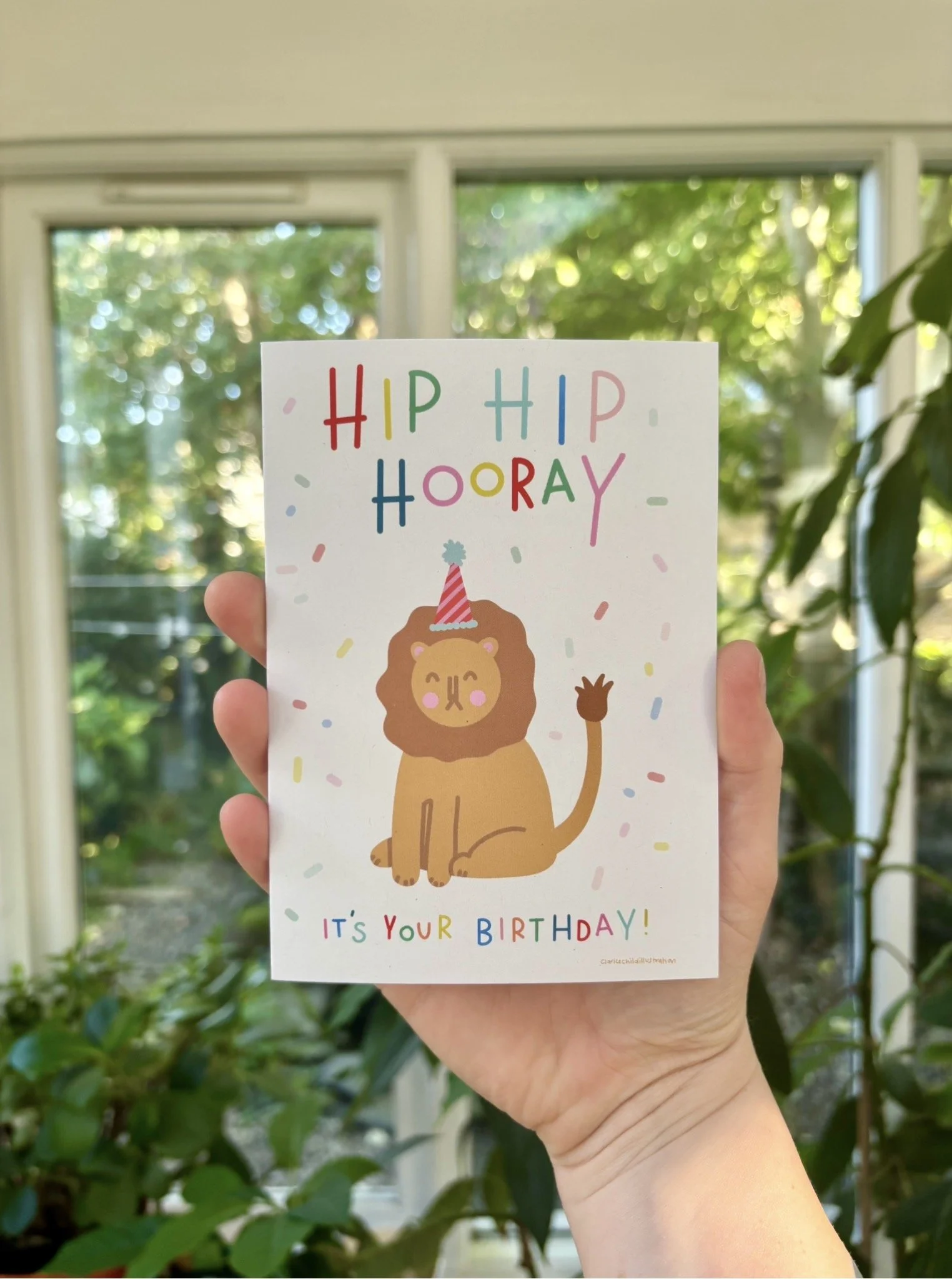 Colorful birthday card with a cartoon lion wearing a party hat, surrounded by confetti, held in front of a window with green trees outside.