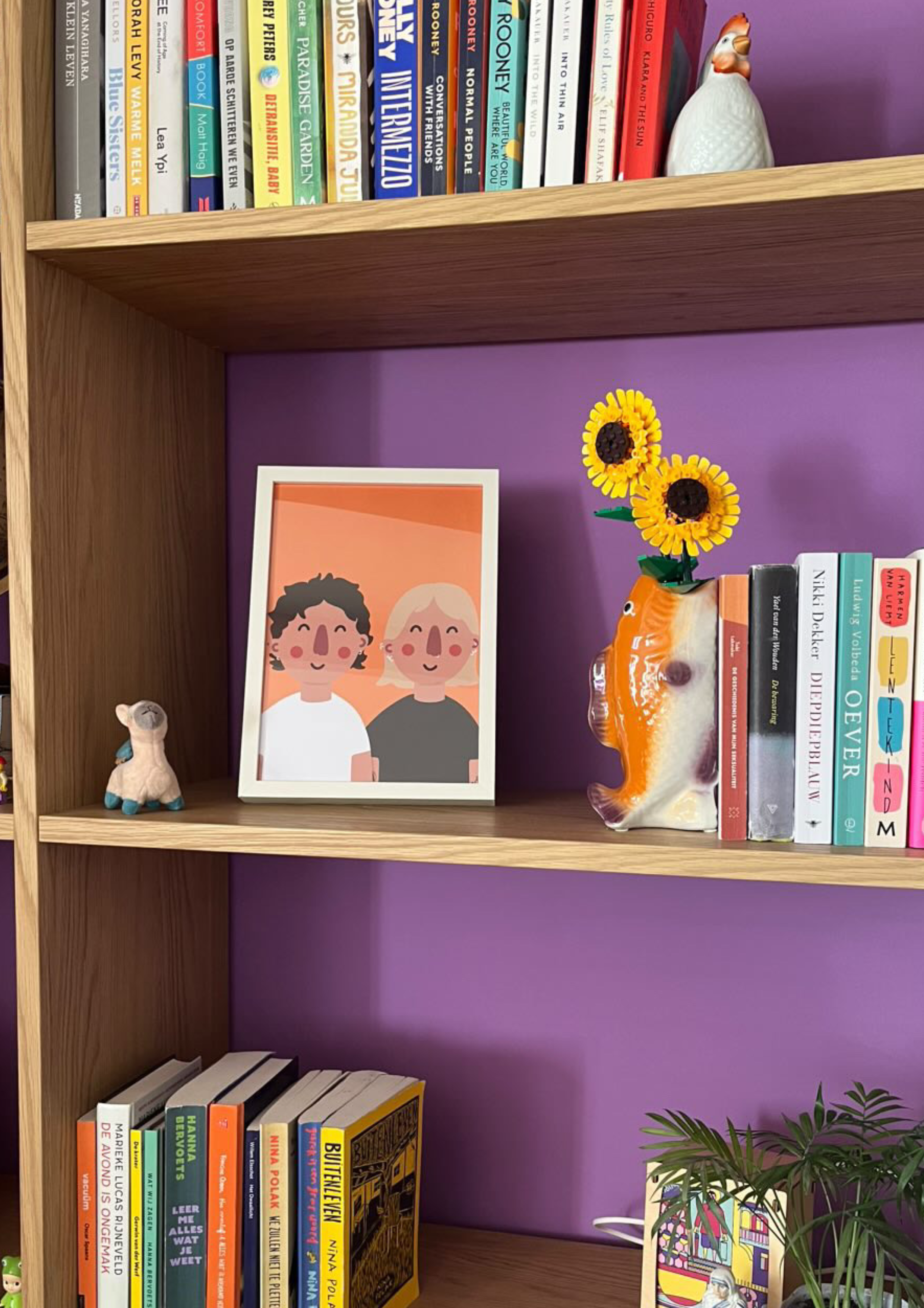 A wooden bookshelf with three shelves, painted purple background. The top shelf has a row of children's and adult books and a decorative ceramic rooster. The middle shelf has a framed cartoon illustration of two smiling characters, a small white llam