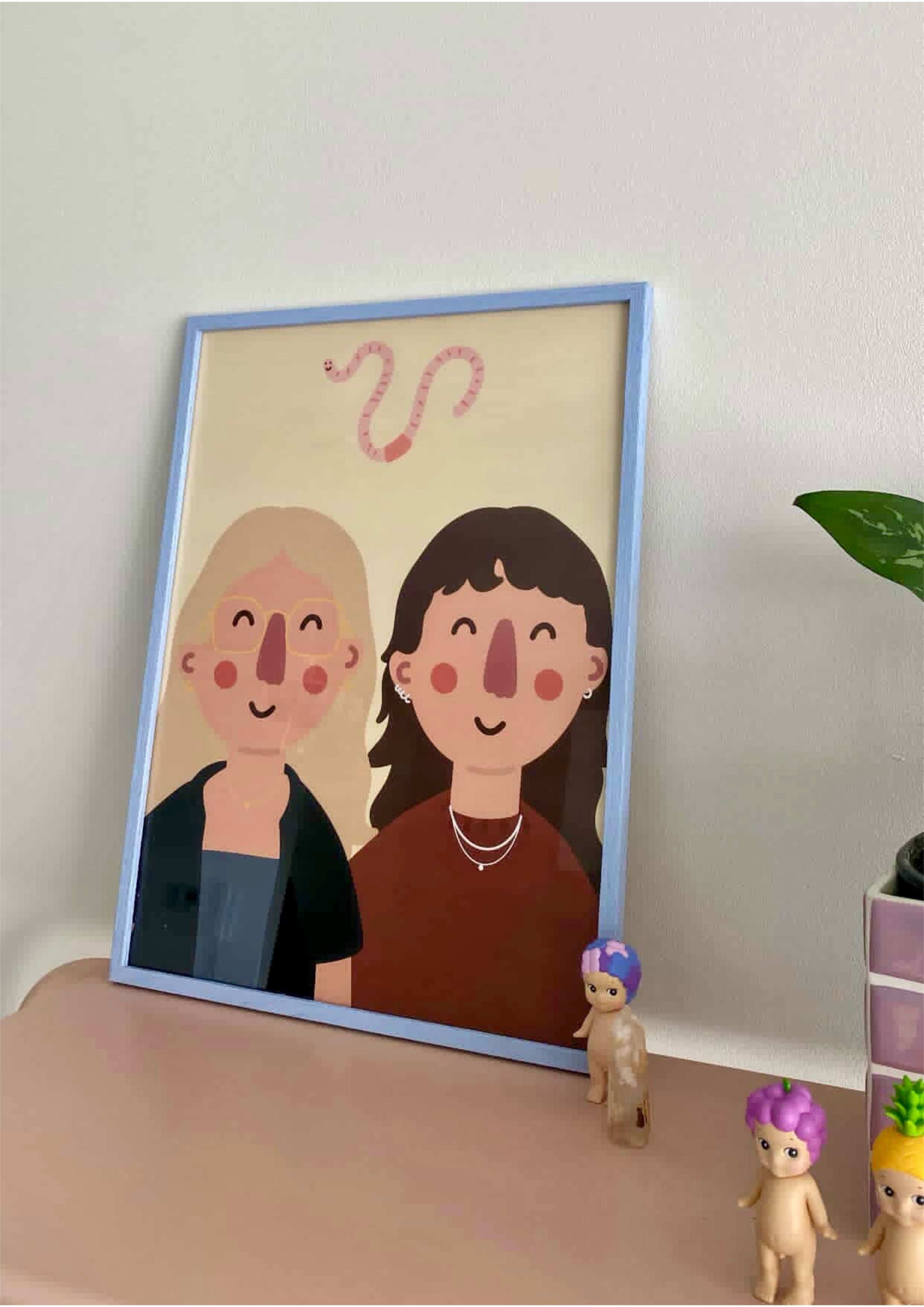A colorful framed illustration of two cartoon women with smiling faces, one with blonde hair and glasses, the other with dark hair and earrings, on a white wall. Small toy dolls with purple and yellow hair, some with animal features, are placed on a 