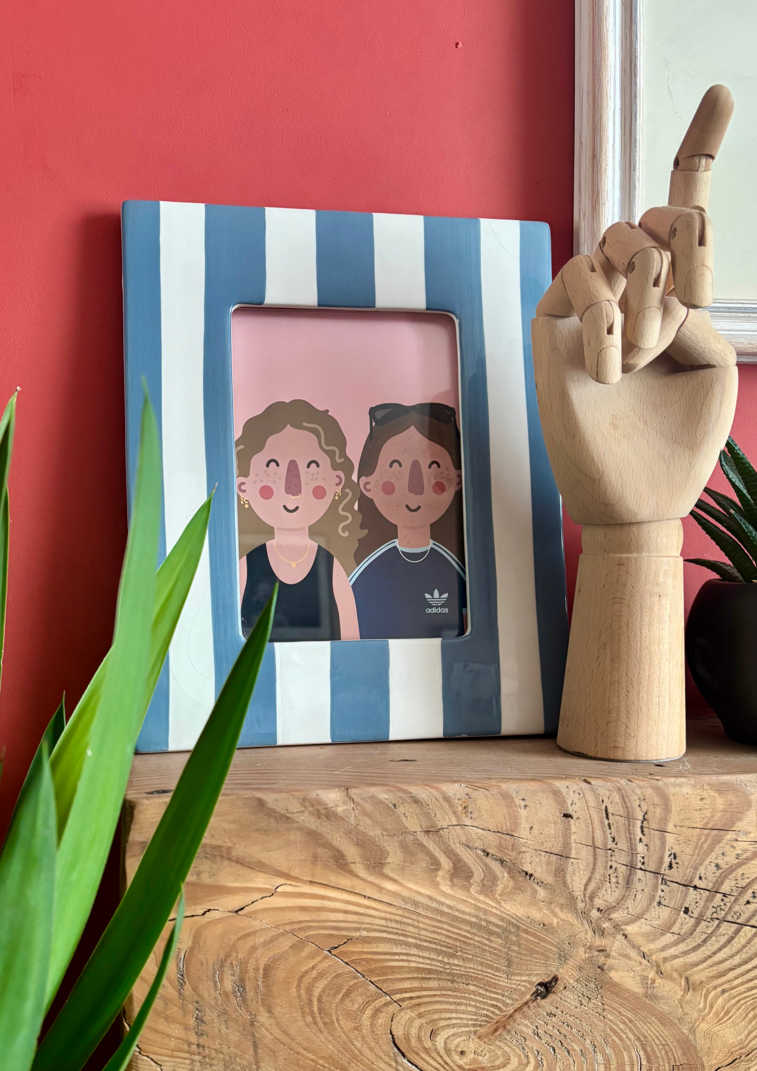 Decorative photo frame with striped blue and white border, featuring car illustration of two smiling women, one with curly hair and earrings, the other with dark hair and sunglasses, placed on a wooden surface next to a wooden mannequin hand and gree