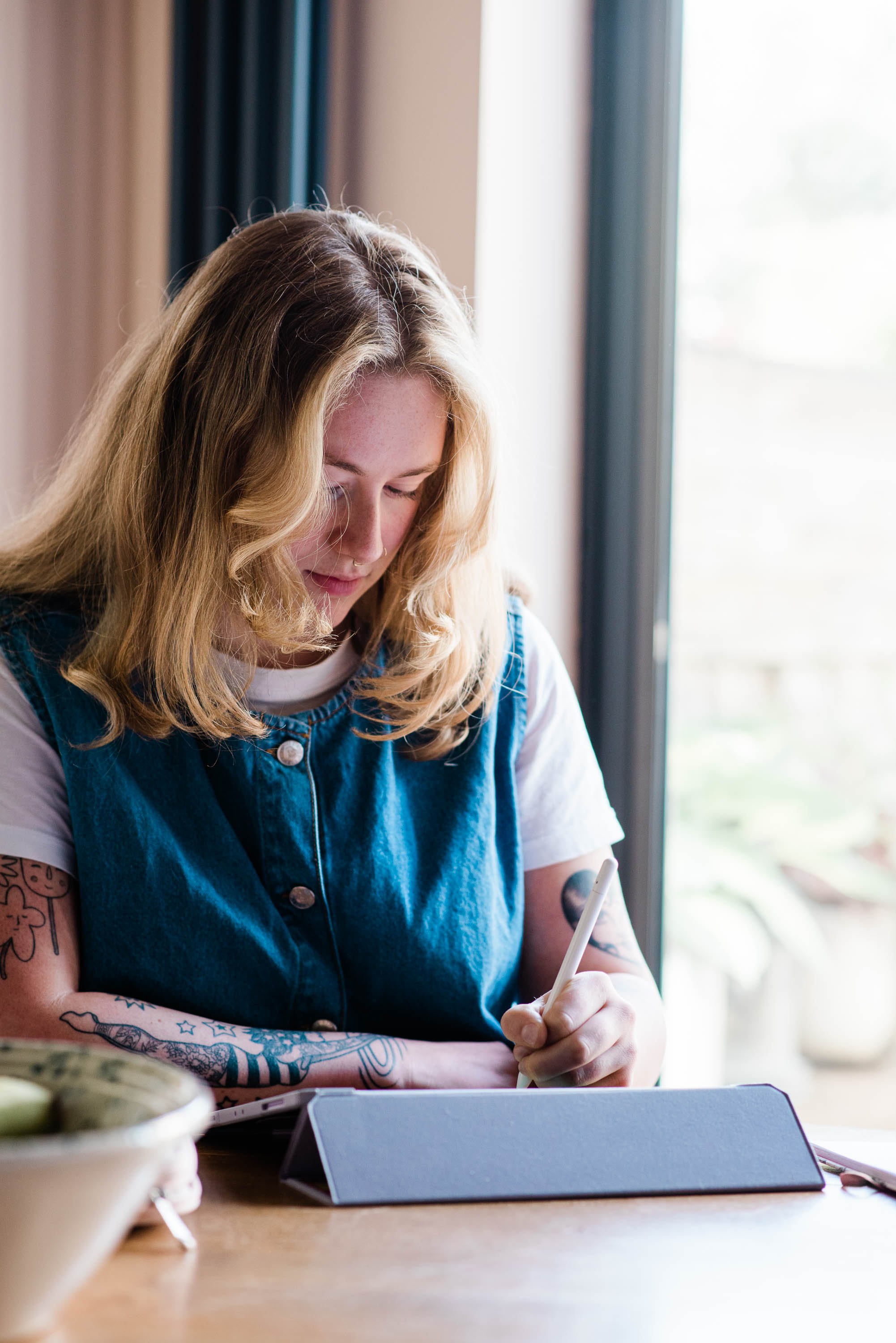 A young woman with long, wavy blonde hair, tattoos on her arms, wearing a denim vest over a white t-shirt, sitting at a wooden table near a window, using a stylus on a tablet.