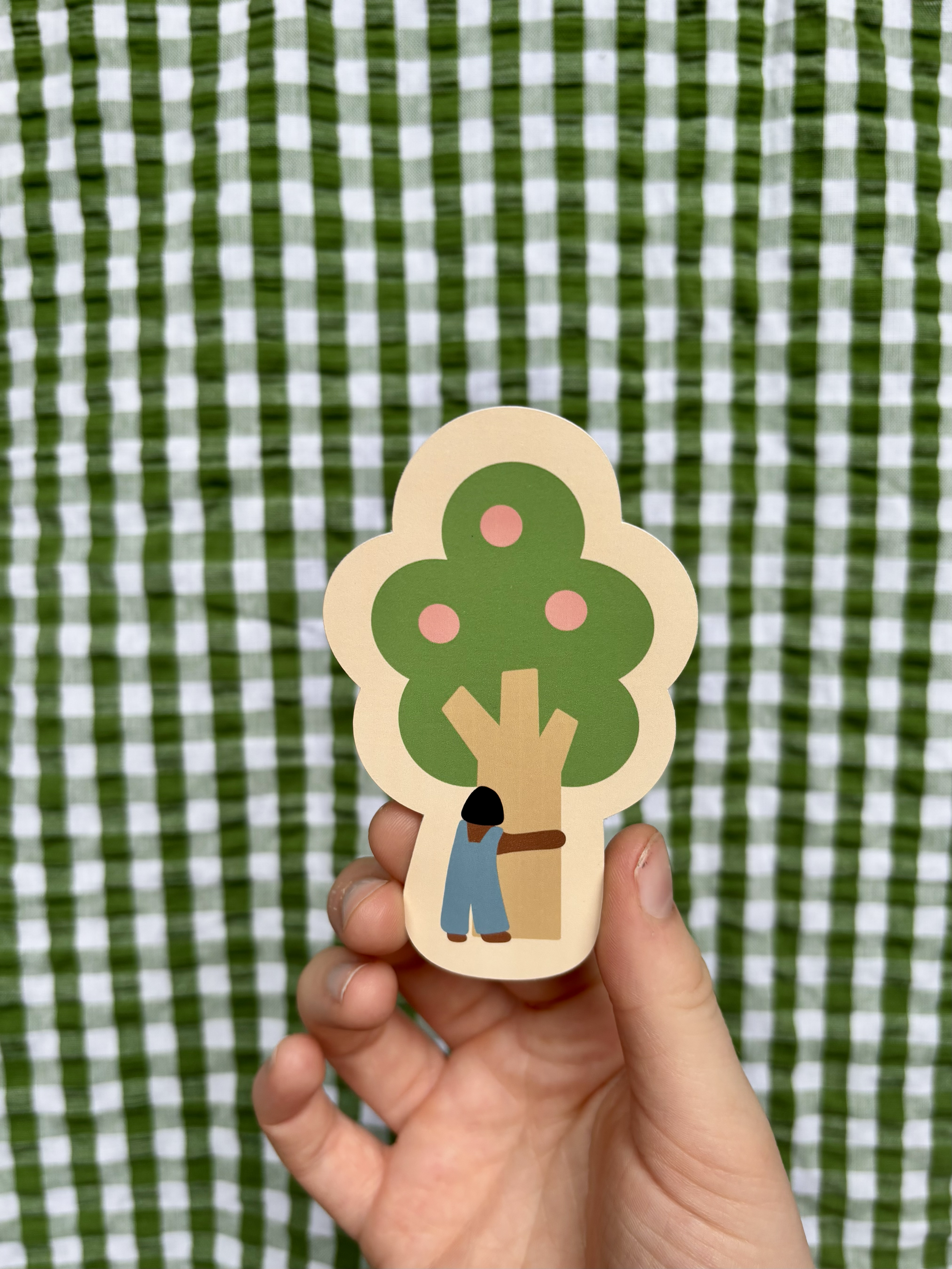 A hand holding a paper cutout of a tree with a person hugging it, set against a green and white checkered background.