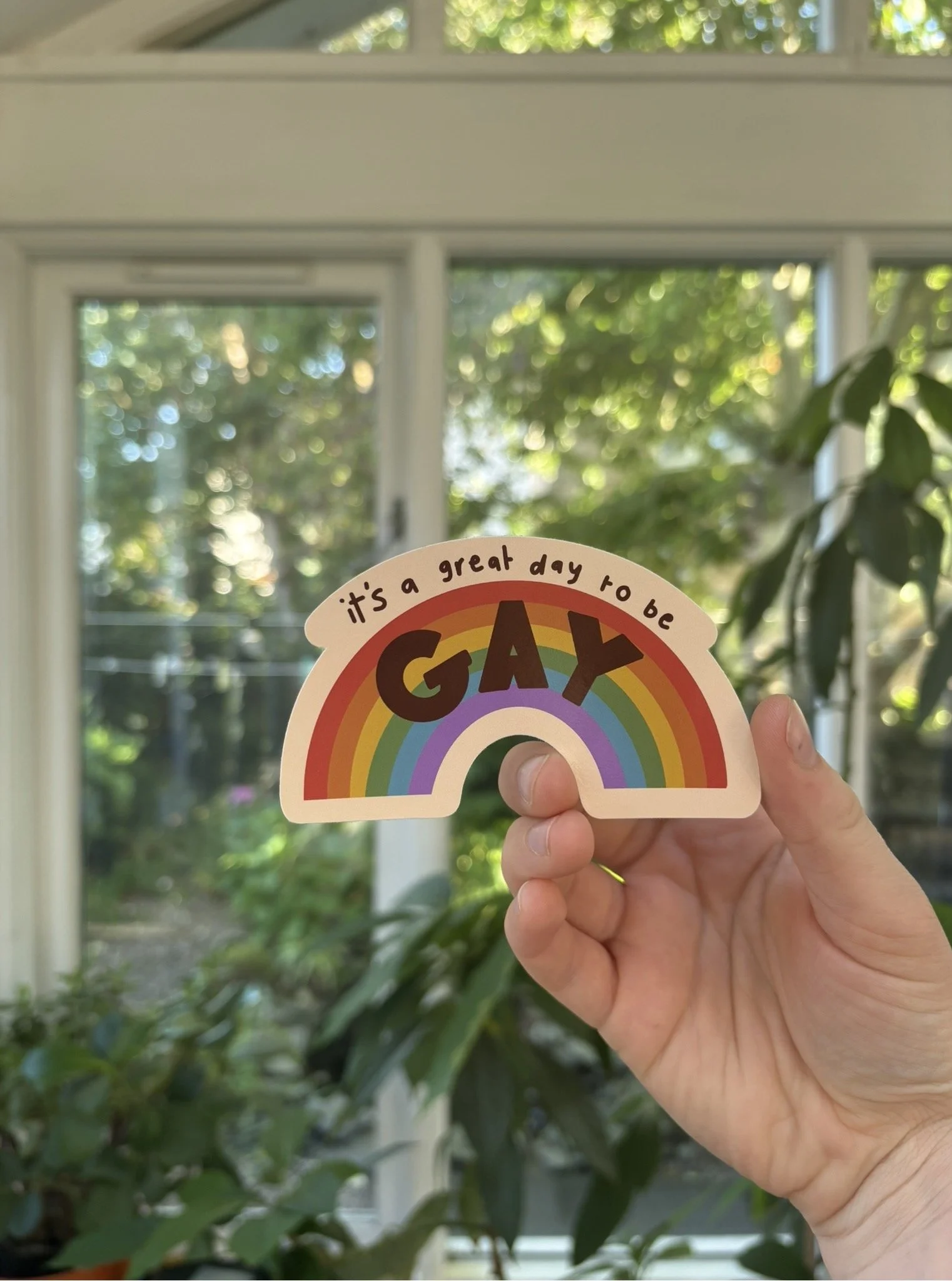 A hand holding a rainbow-colored sticker with the words 'it's a great day to be happy' on a bright, sunny day. In the background, a window with a view of green foliage.