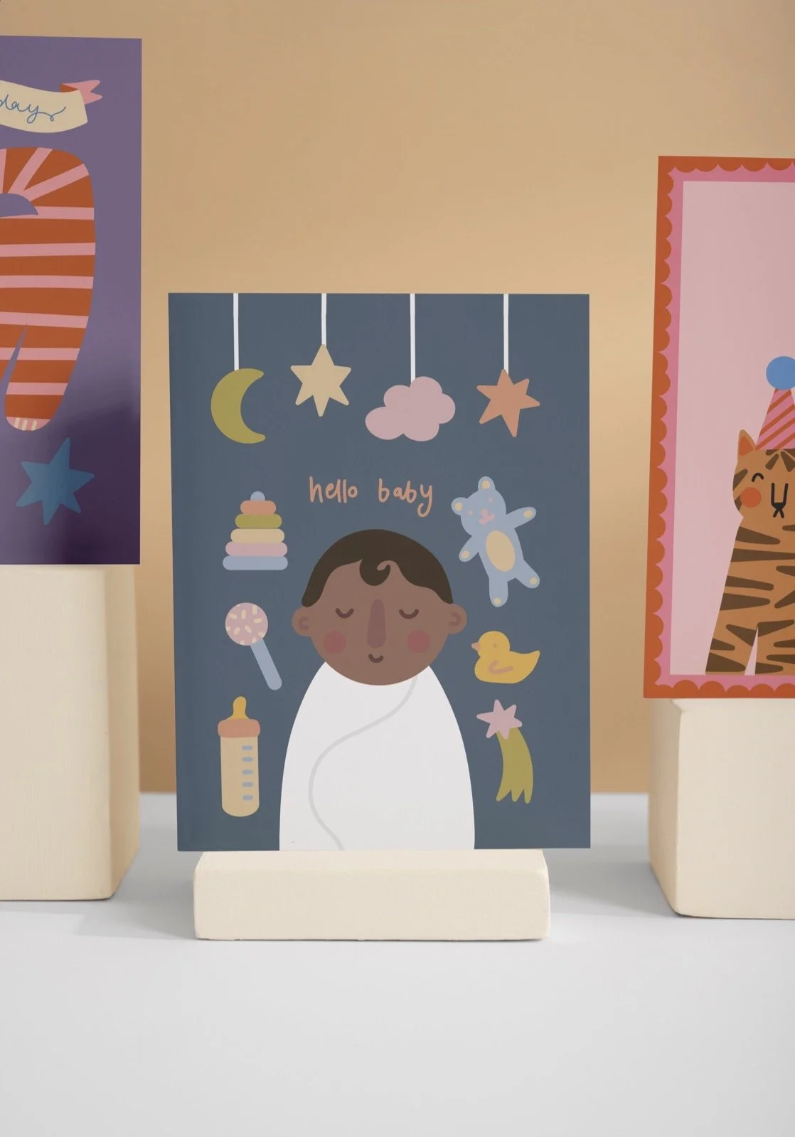 Colorful baby shower or nursery card with a cartoon baby wrapped in a blanket, surrounded by baby-related items like a rattle, bottle, rubber duck, stacking rings, and teddy bear, with moon and star decorations, and the words "hello baby".