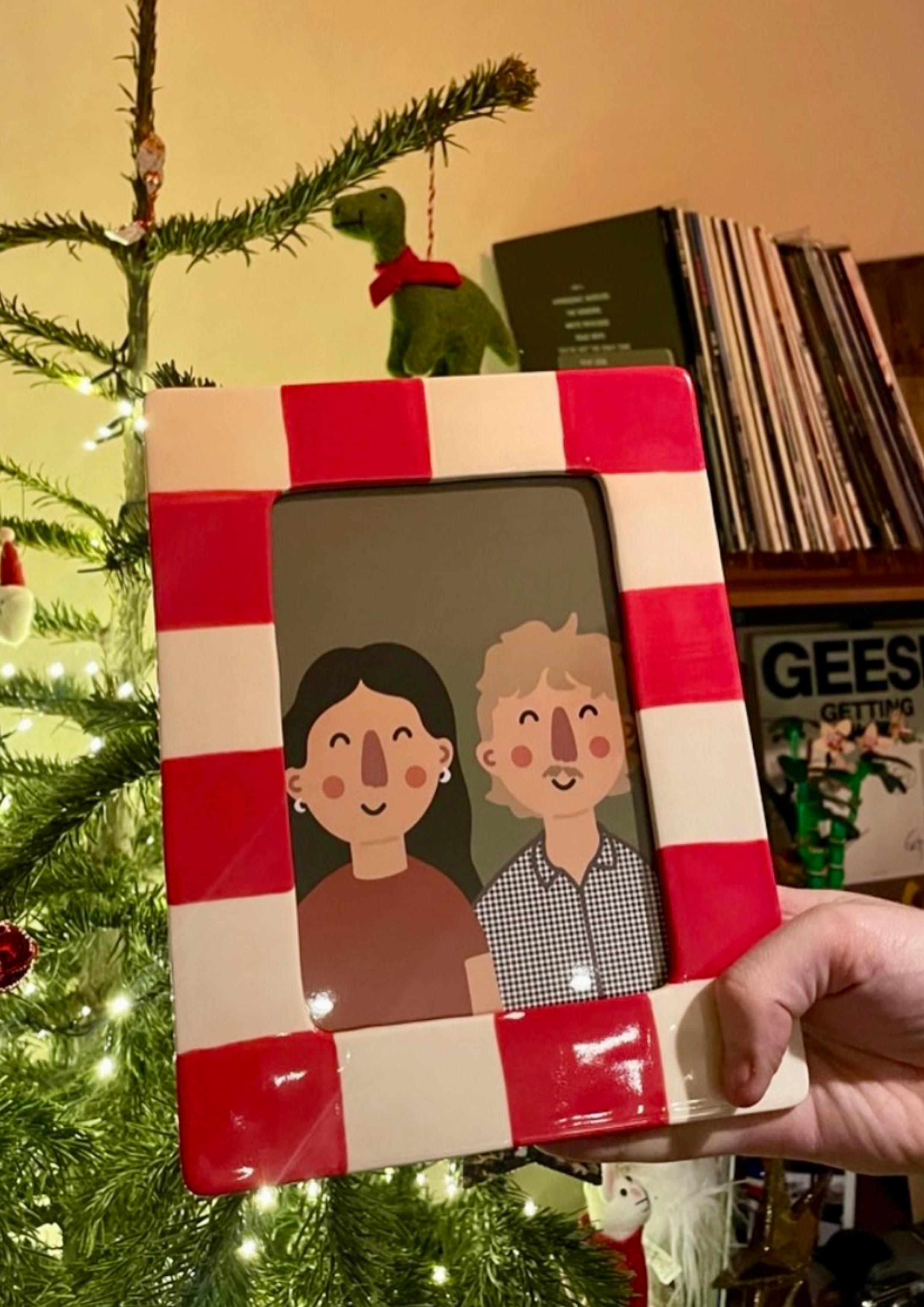 A hand holding a framed cartoon illustration of a smiling woman with dark hair and a smiling man with blond hair and a checkered shirt. The frame is decorated with red and white stripes and is placed next to a Christmas tree with lights and ornaments