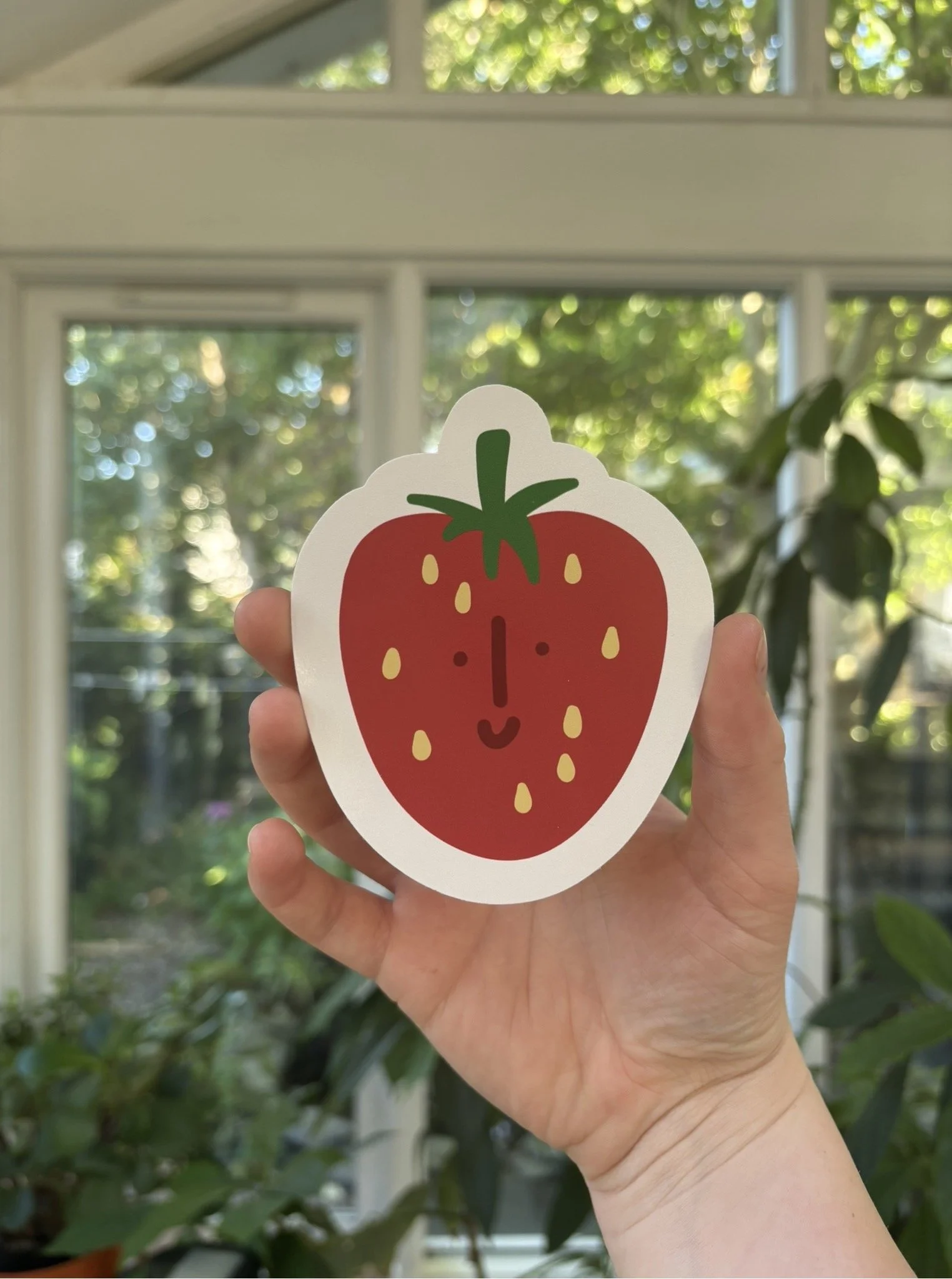 A hand holding a sticker of a smiling cartoon strawberry with seeds, green top, and a happy face, against a background of a window with green leaves outside.