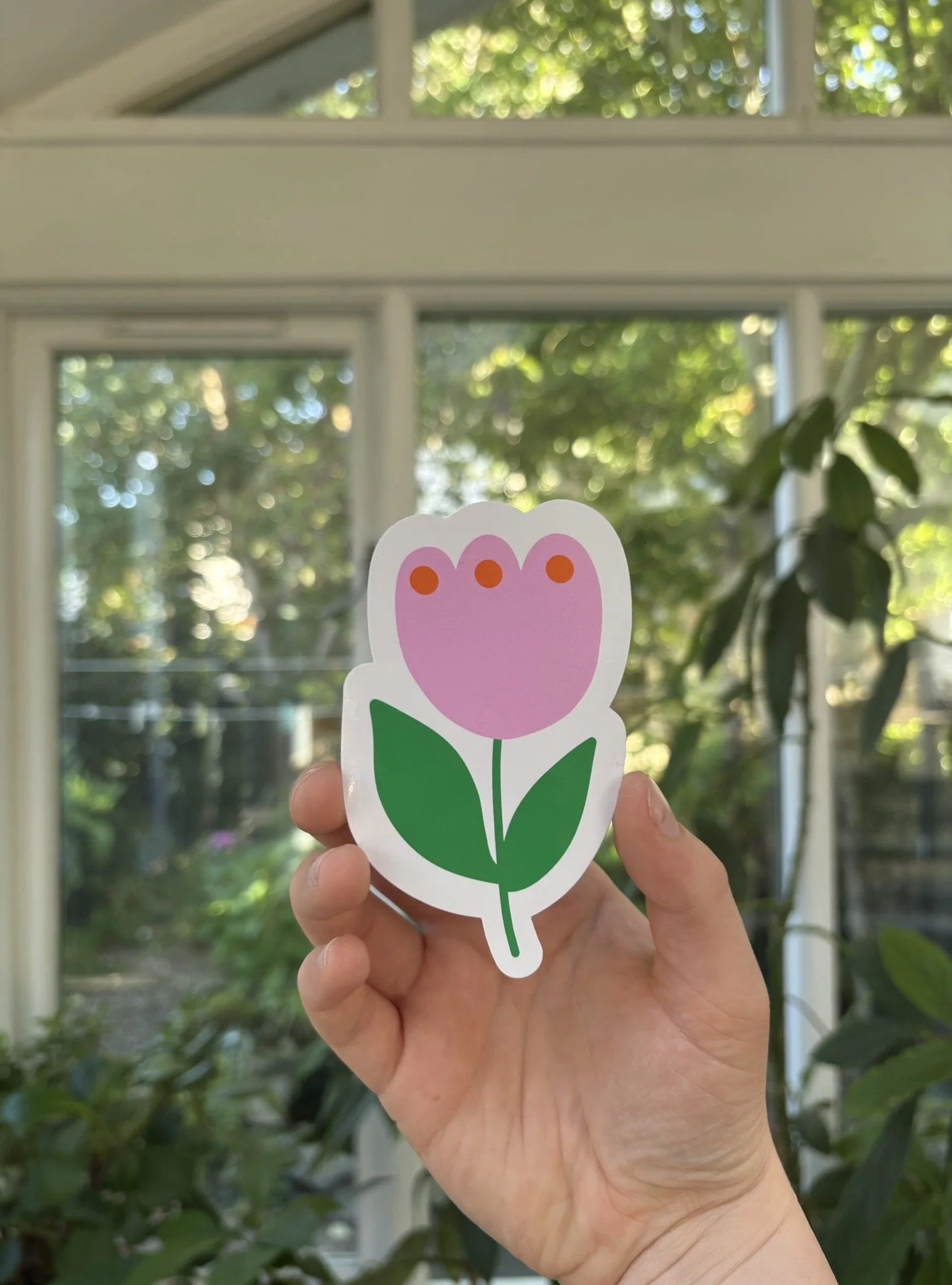 Hand holding a paper cutout of a pink flower with green leaves in front of a window and greenery outside.