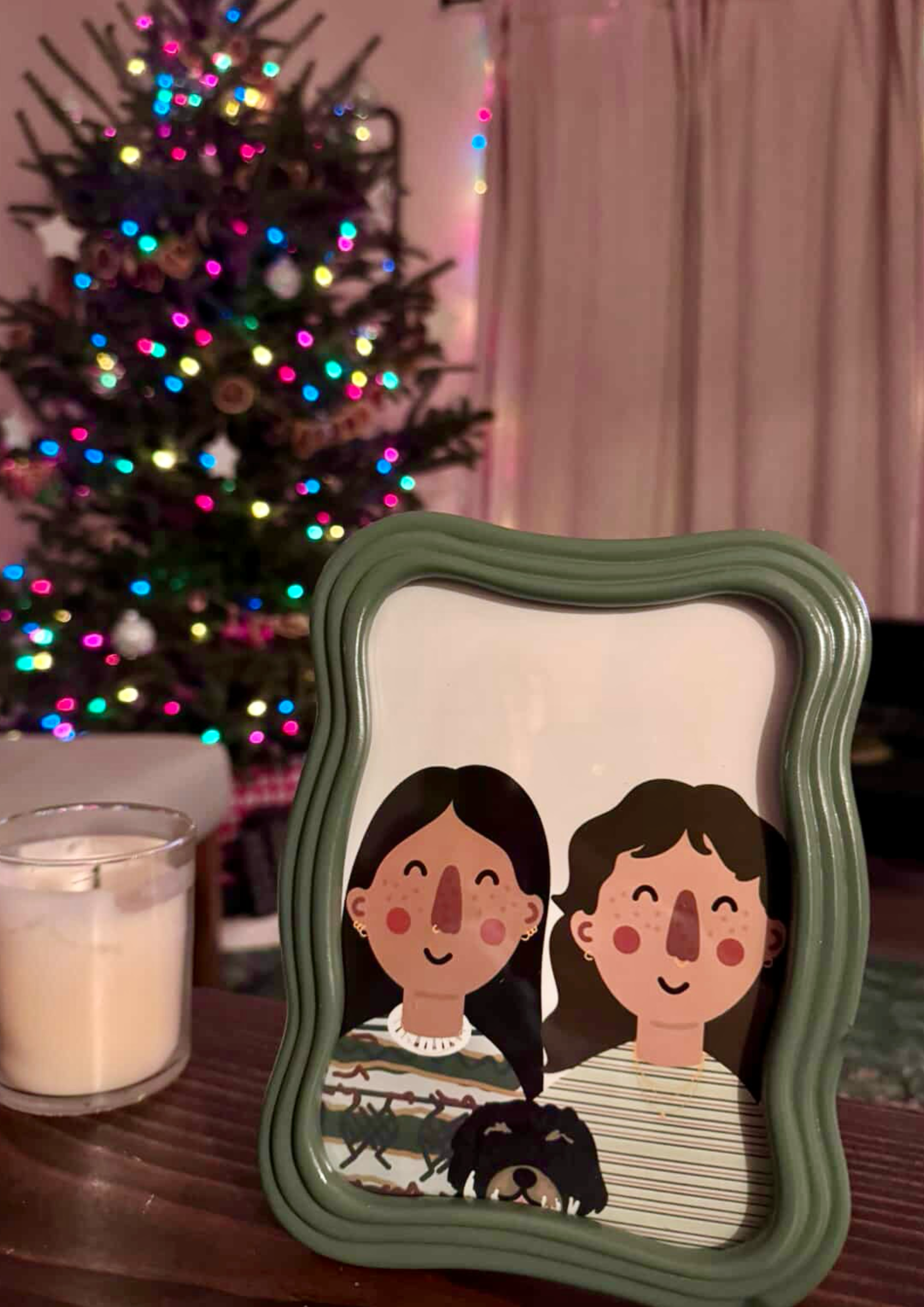 A green-framed mirror with cartoon illustration of two smiling women with brown and black hair, set on a wooden table. Behind the mirror, there is a Christmas tree decorated with colorful lights and ornaments. A candle and a striped box with a dog il