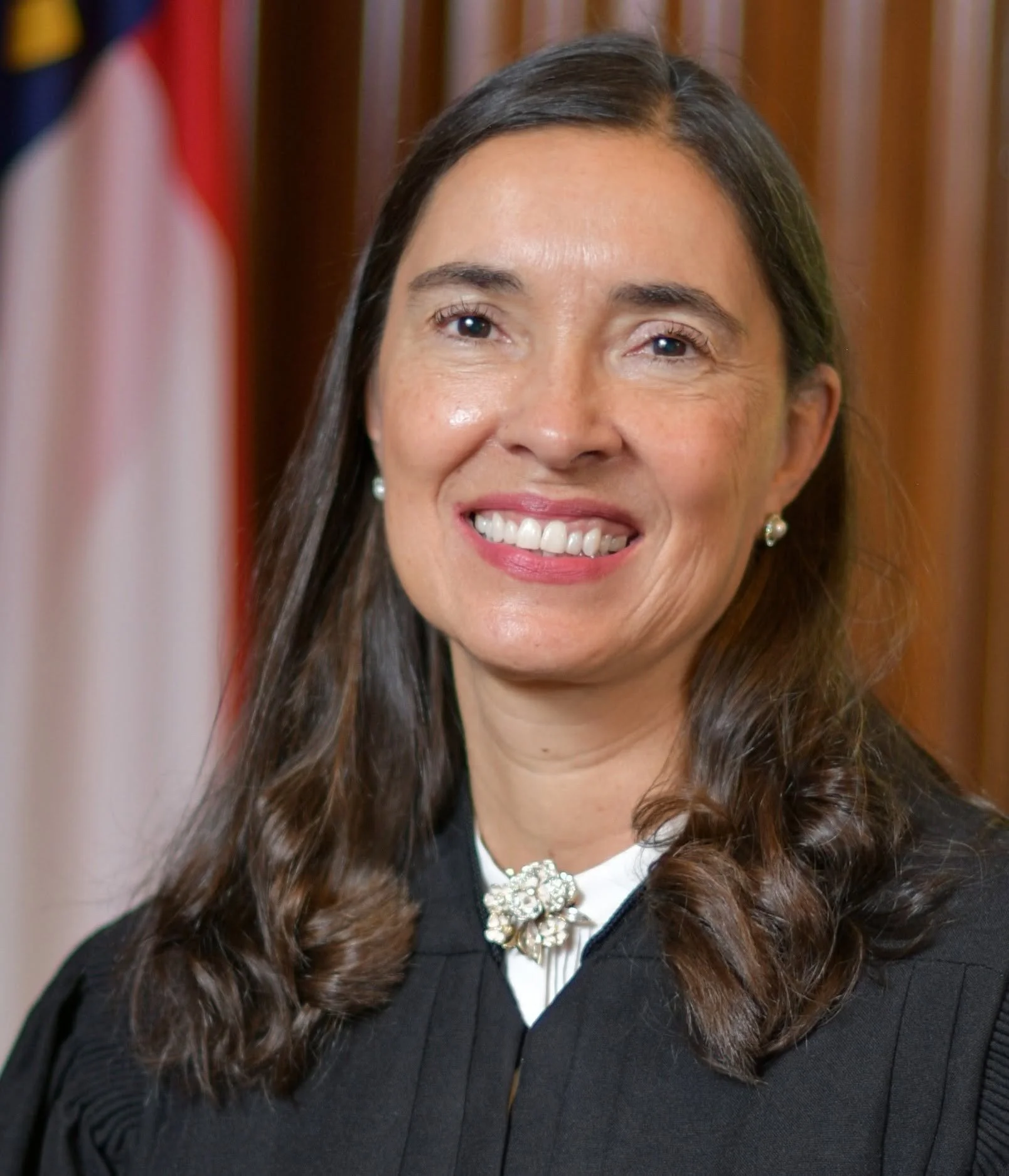 A woman with long brown hair smiling, wearing pearl earrings, a black blazer, and a white blouse with a decorative brooch, standing in front of a wooden background and a blurry American flag.