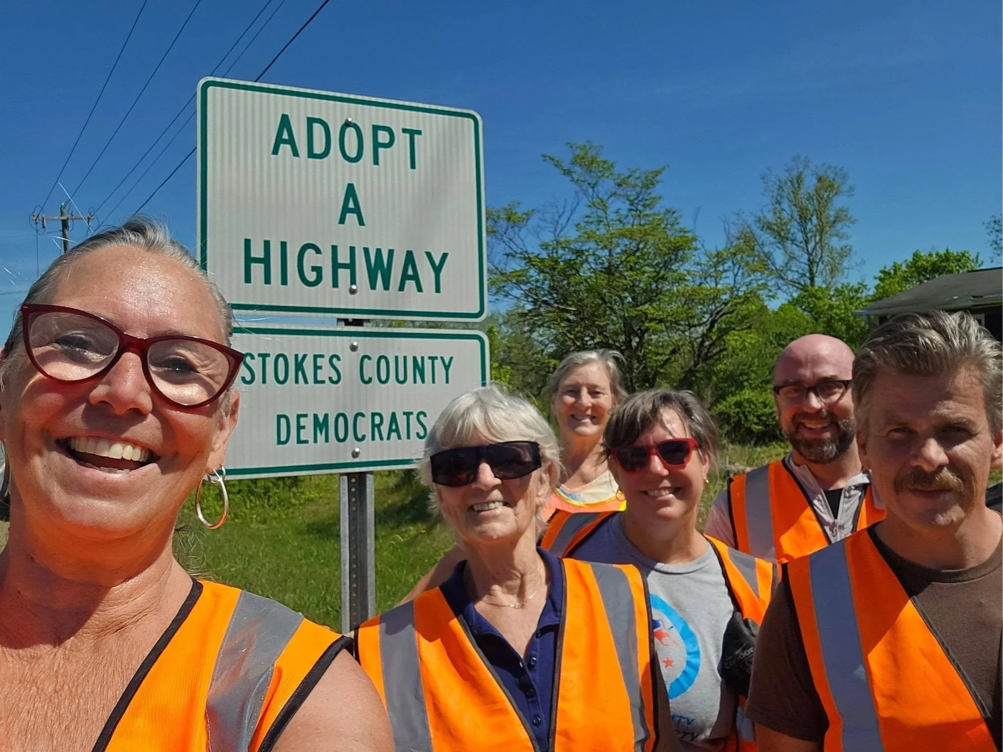 A group of smiling people wearing orange safety vests taking a group selfie outdoors near a highway sign that reads 'Adopt A Highway, Stokes County, Democrats.' The sky is clear and there are trees in the background.