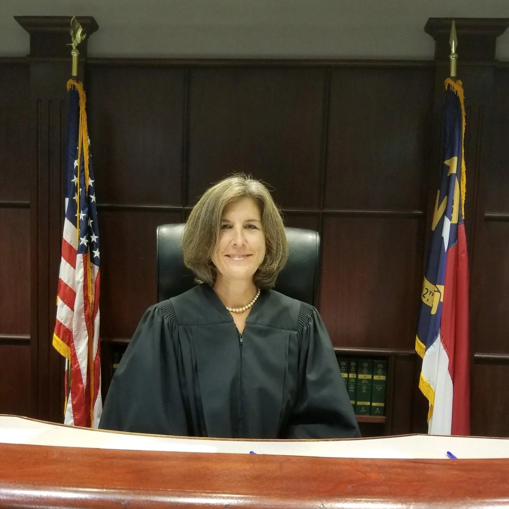 A woman sitting in a courthouse or legal office behind a wooden desk, wearing a judge's robe, with the American flag and another official flag behind her, dark wood paneling, and legal books.