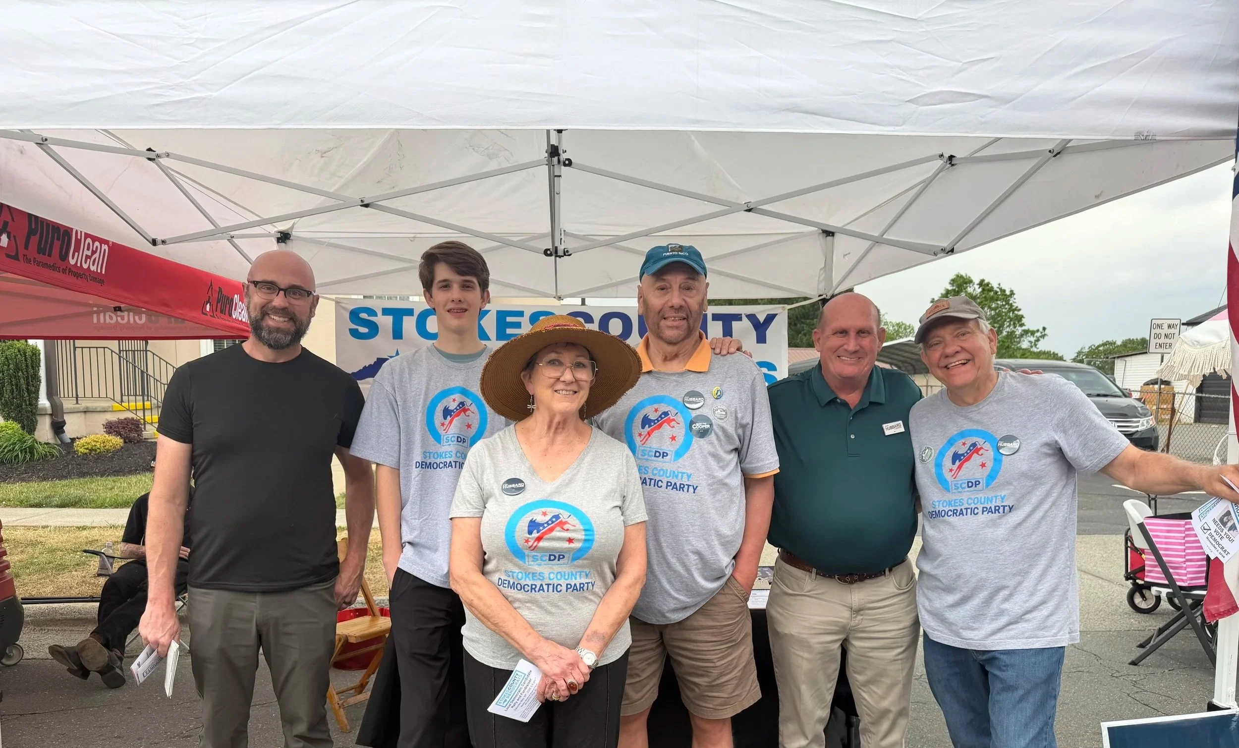 A group of six people standing together outdoors under a white canopy tent at a community event, wearing T-shirts with the Stokes County Democratic Party logo, smiling for the camera. There is a banner behind them that reads 'STOKES COUNTY.'