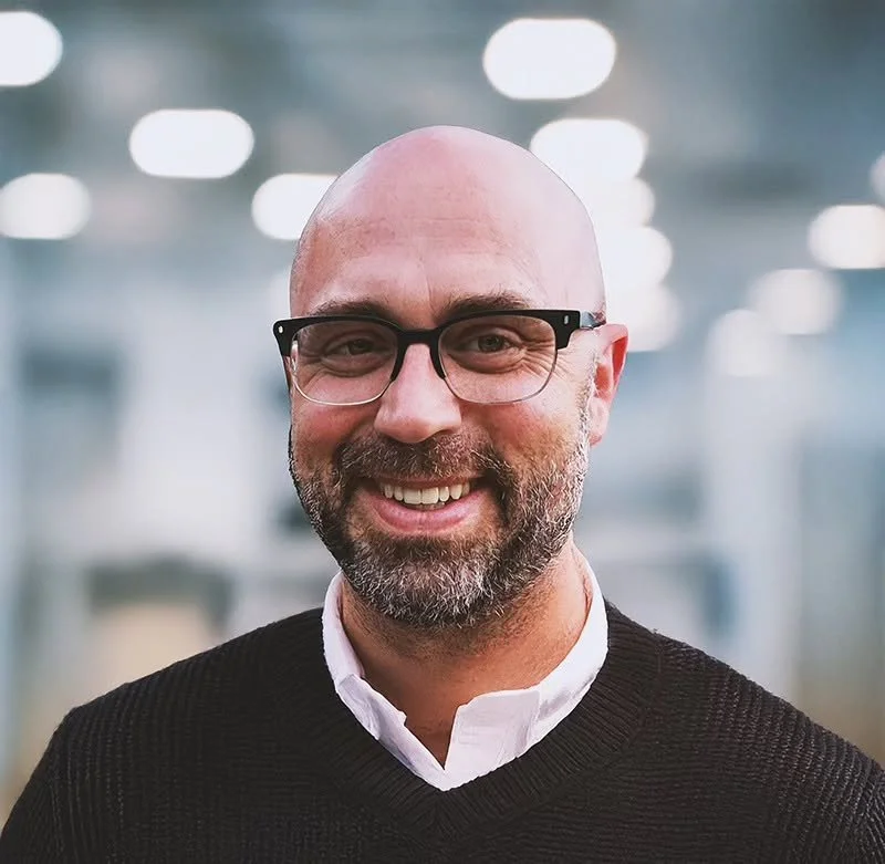 A middle-aged man with glasses, a beard, and a big smile, wearing a white shirt and a black sweater, standing in an indoor setting with blurred lights in the background.