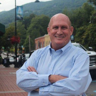 A smiling bald man in a light blue dress shirt with arms crossed standing outdoors on a city street with trees, buildings, and cars in the background.