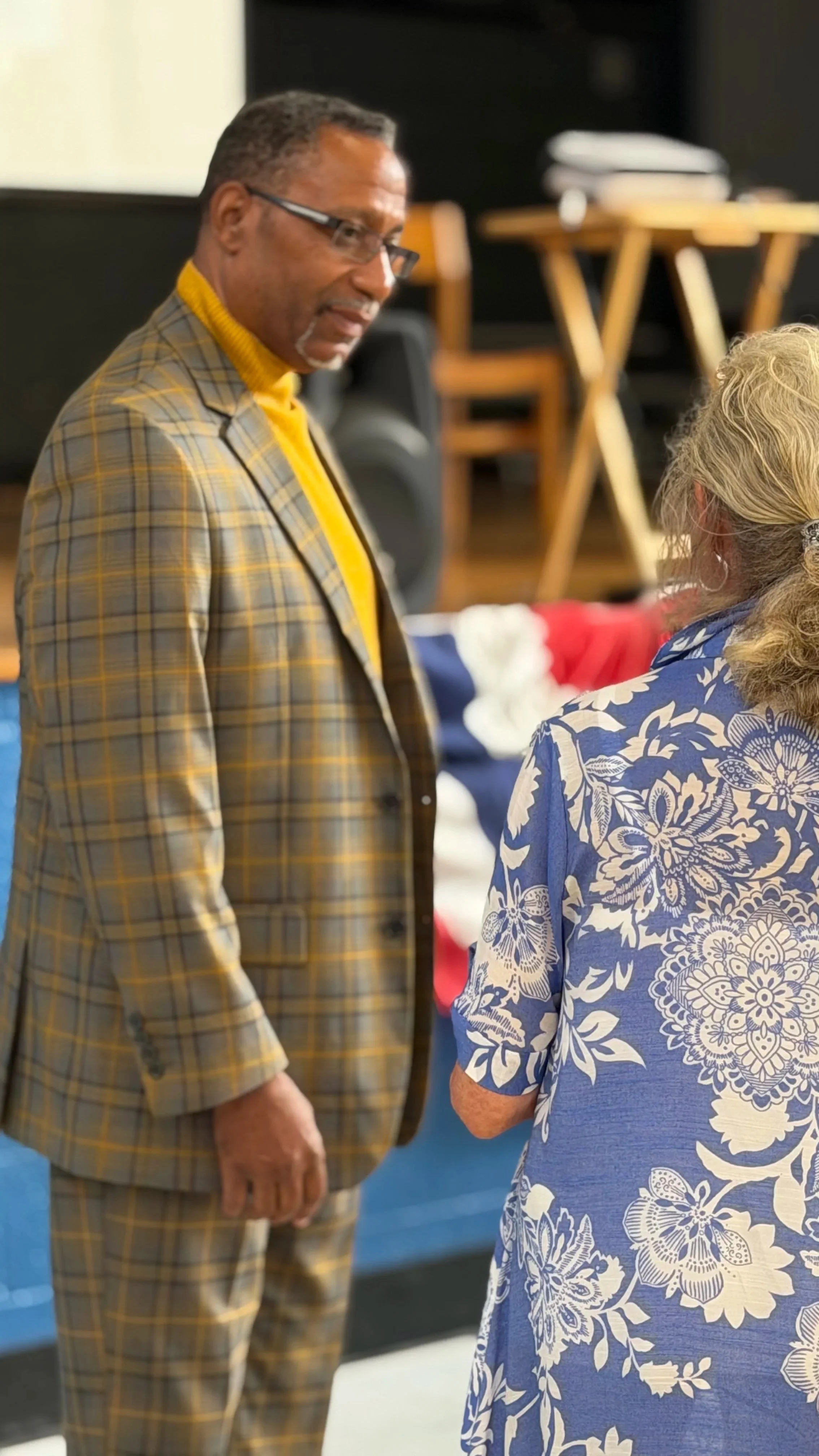 A man and a woman speaking to each other indoors, with tables and chairs in the background. The man is wearing glasses, a yellow turtleneck, and a plaid suit. The woman has blonde hair and is wearing a blue dress with white floral patterns.
