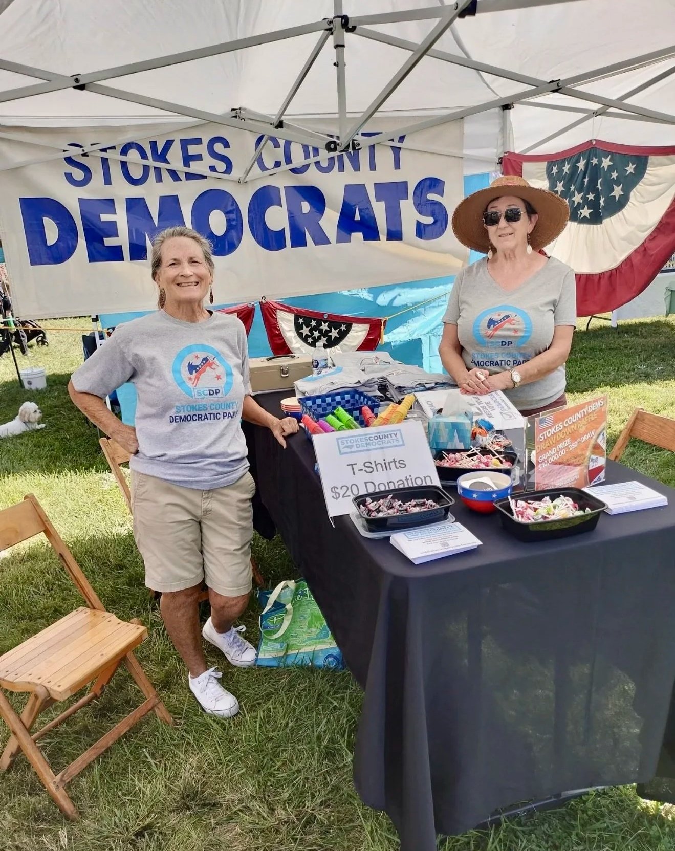 Two women standing at a booth for Stokes County Democrats, with a banner reading 'Stokes County Democrats' behind them. The booth offers T-shirts for $20 donation and various stickers and buttons. The women are smiling, one wearing khaki shorts and a gray T-shirt, the other in a straw hat, sunglasses, and a gray T-shirt. There are American-themed decorations and informational materials on the table.