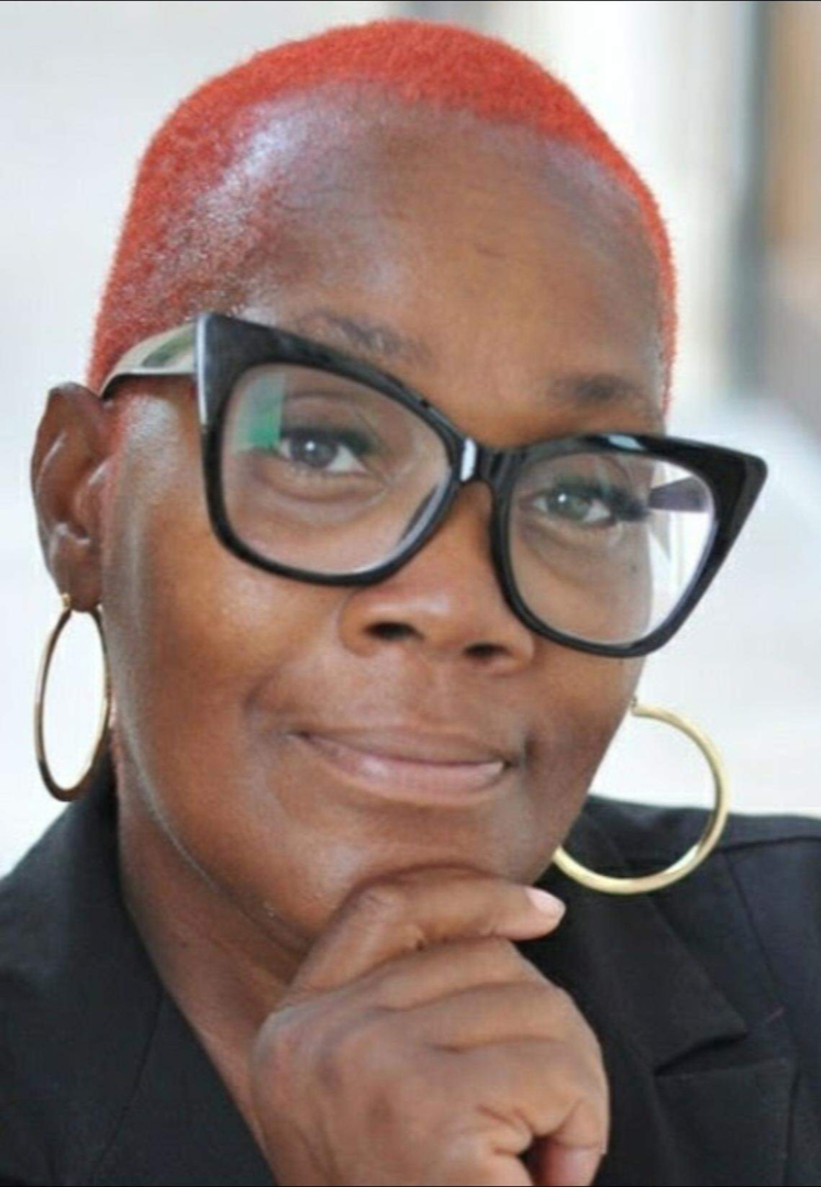 A woman with short, bright red hair wearing large black glasses and gold hoop earrings, smiling softly and resting her chin on her hand.