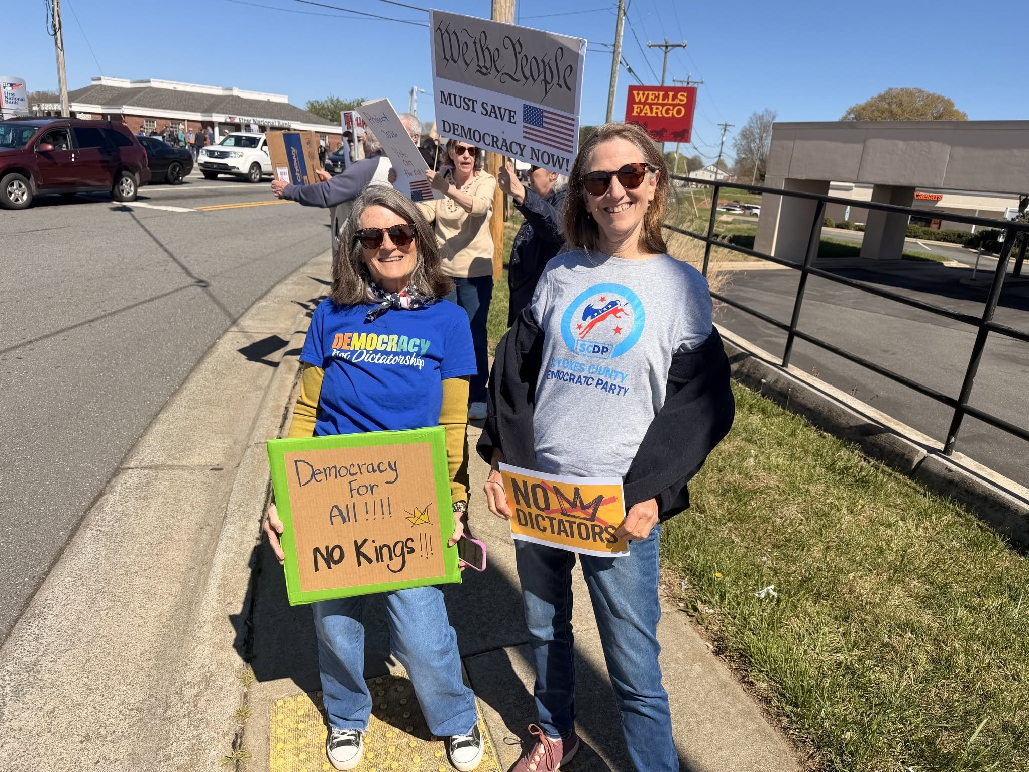 Two women smiling and holding protest signs on a sidewalk during a political protest. The sign on the left reads, "Democracy for All! No Kings! No Dictators!" The sign on the right reads, "We the People Must Save Democracy Now!" In the background, multiple people are holding signs including one that says, "Project Vote" and another with the Wells Fargo logo. Cars are parked along the street, and a bank building and a restaurant are visible in the background.