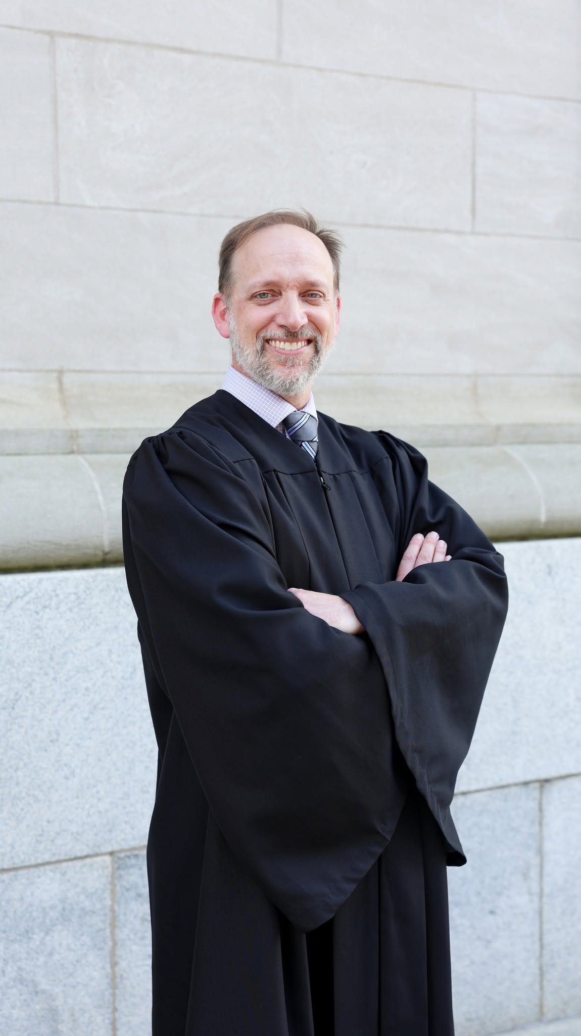 A man in a graduation gown standing outside in front of a stone wall, smiling with arms crossed.