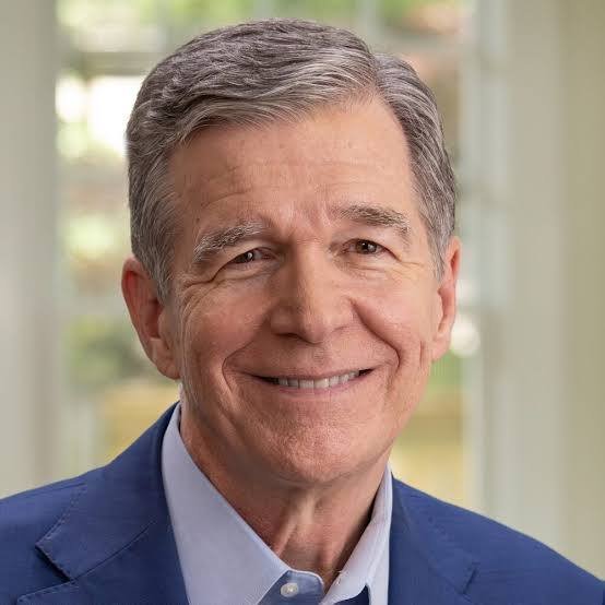 Close-up of a smiling middle-aged man with gray hair, wearing a blue blazer and light blue shirt, indoors with a blurred window background.