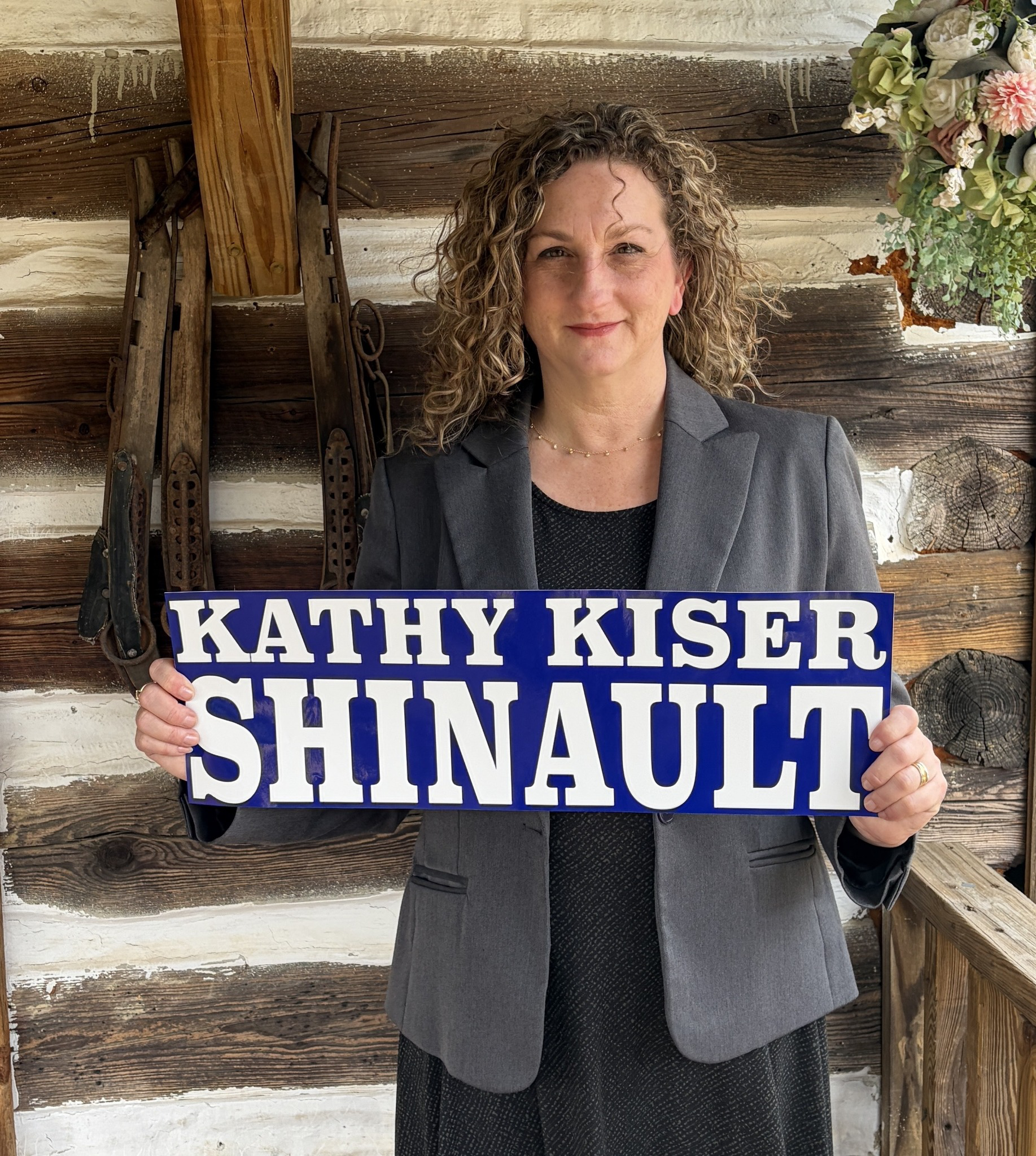 A woman with curly hair and a gray blazer holding a campaign sign that reads 'KATHY KISER SHINAUT' standing in front of a rustic wooden wall with a vintage wooden snowshoe and a bouquet of flowers in the upper right corner.