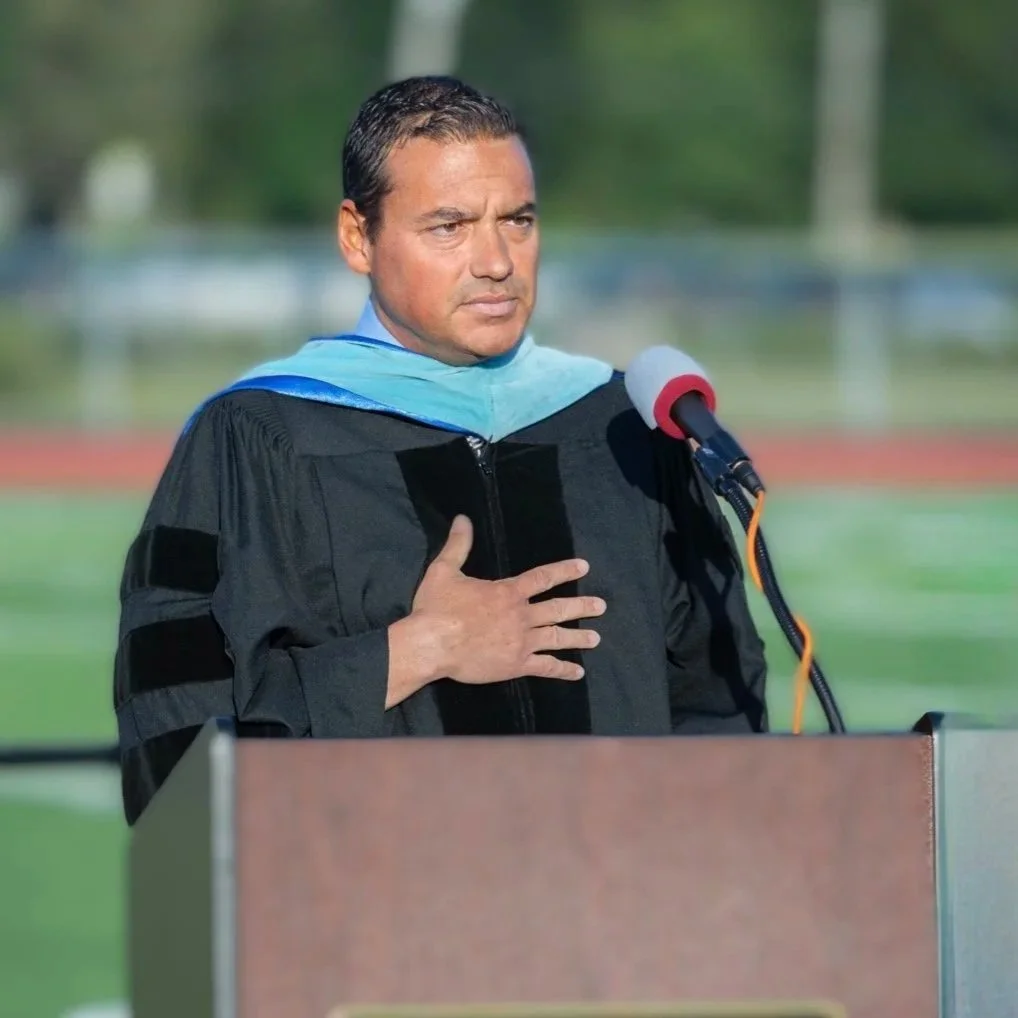 A man in a graduation gown standing at a podium with one hand over his heart, outdoors with a blurred background of trees and sports field.