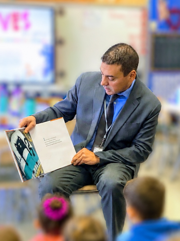 Man in a suit reading a children's book to students in a classroom.