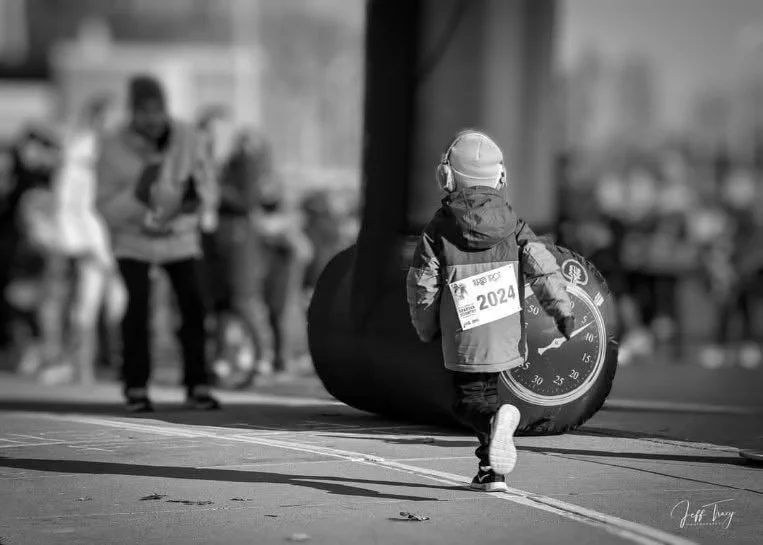 Child running with headphones on, wearing a jacket with a race bib for a 2024 event, in front of a large inflatable clock, with blurred people in the background.
