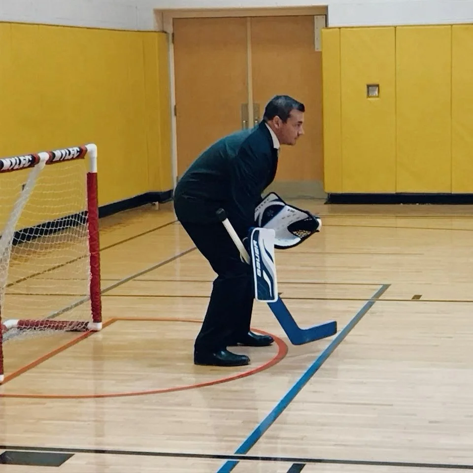 A man dressed in formal clothing, wearing skates and goalie gear, standing in a gymnasium near a hockey goal, preparing to play hockey.