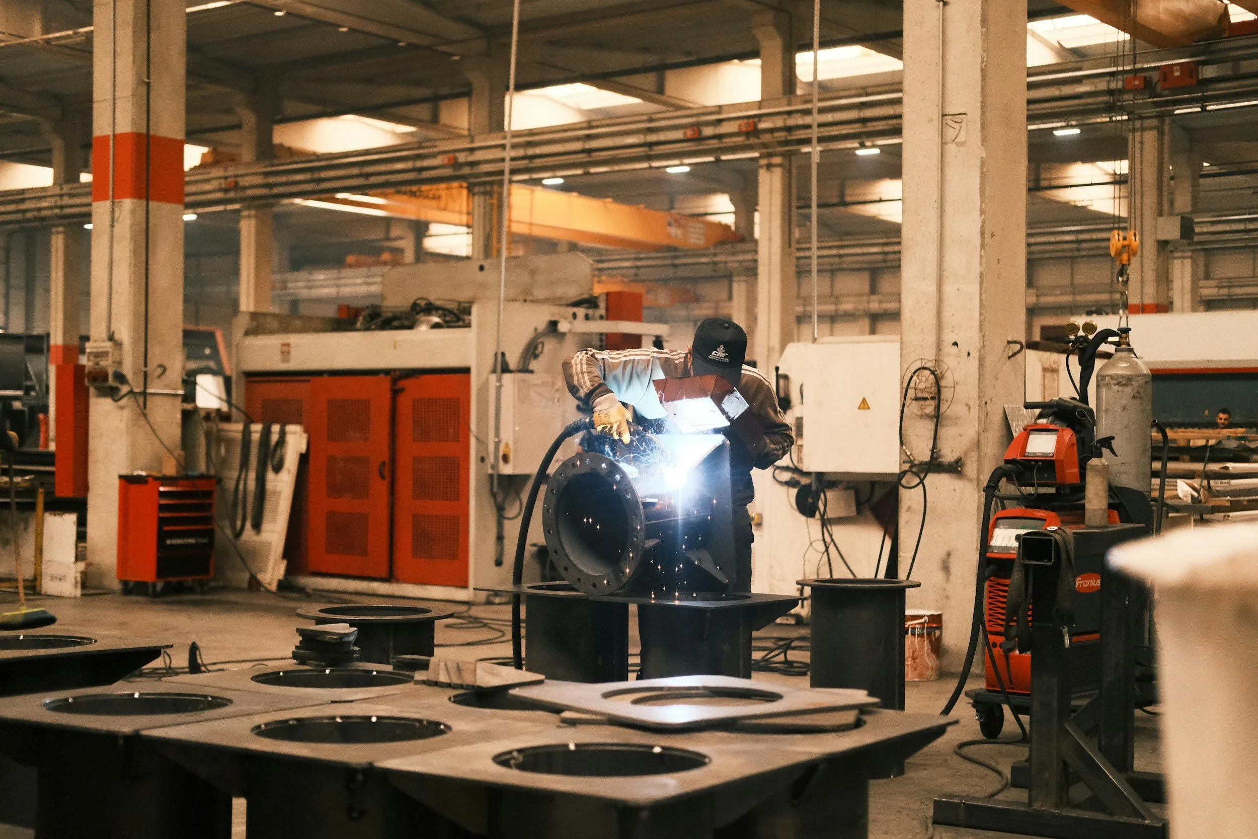 A worker welding a metal pipe in an industrial workshop with machinery and metal components around.