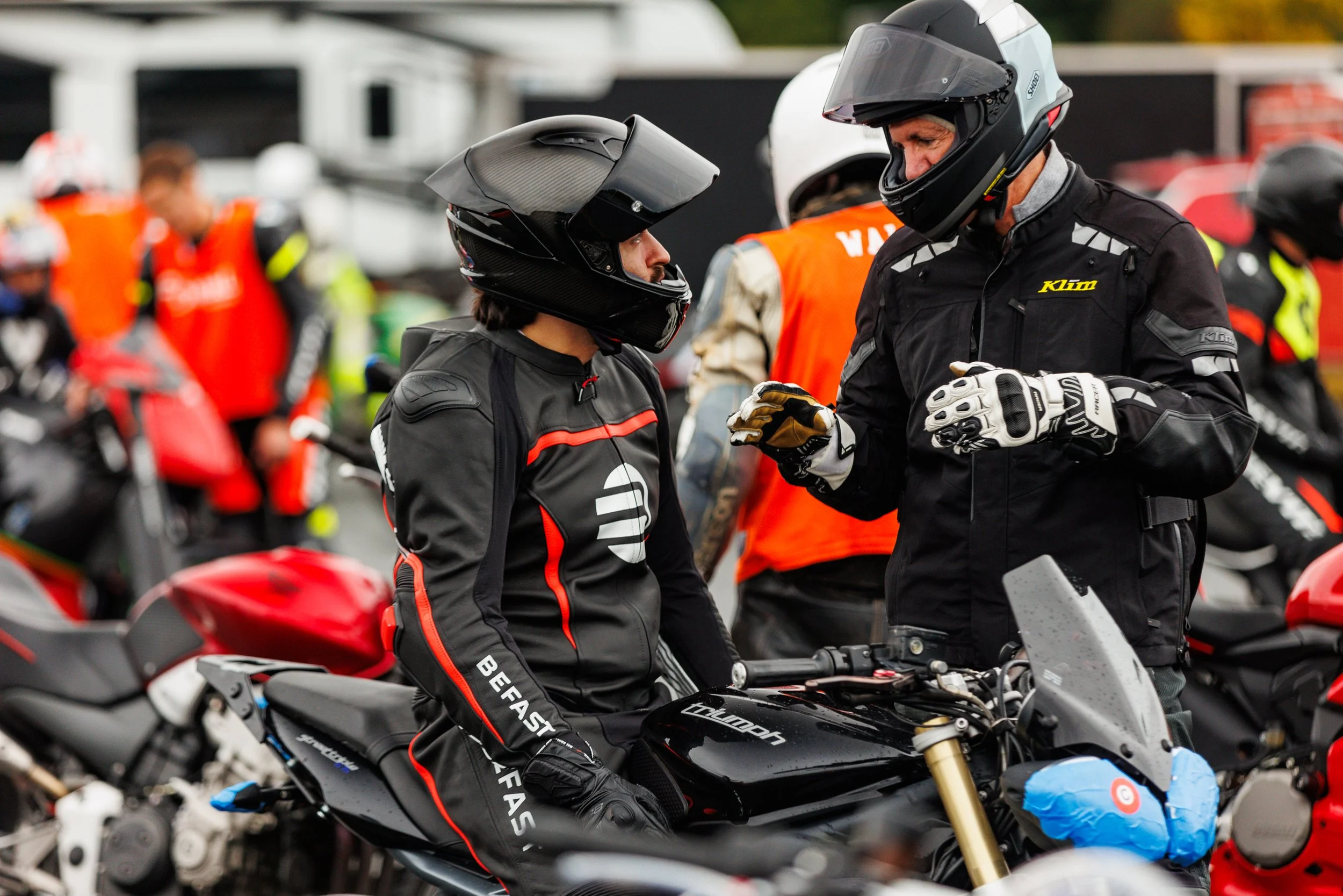 Two motorcyclists in racing gear and helmets talking among a group of riders and motorcycles at a racing event.