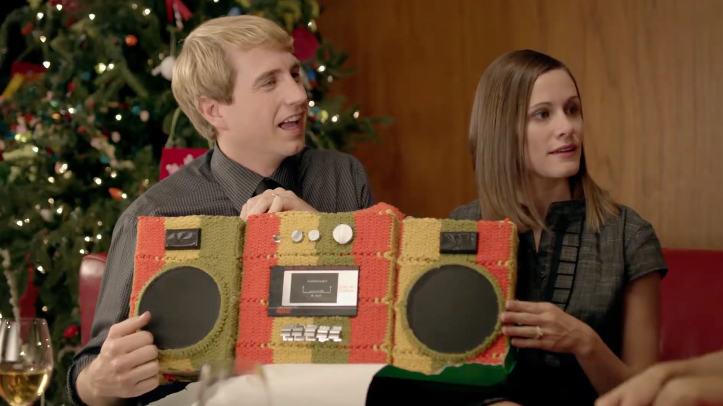 A young man and woman are sitting at a table during a holiday celebration. The man is holding a large, colorful boombox made from a knitted material, and there is a decorated Christmas tree in the background.