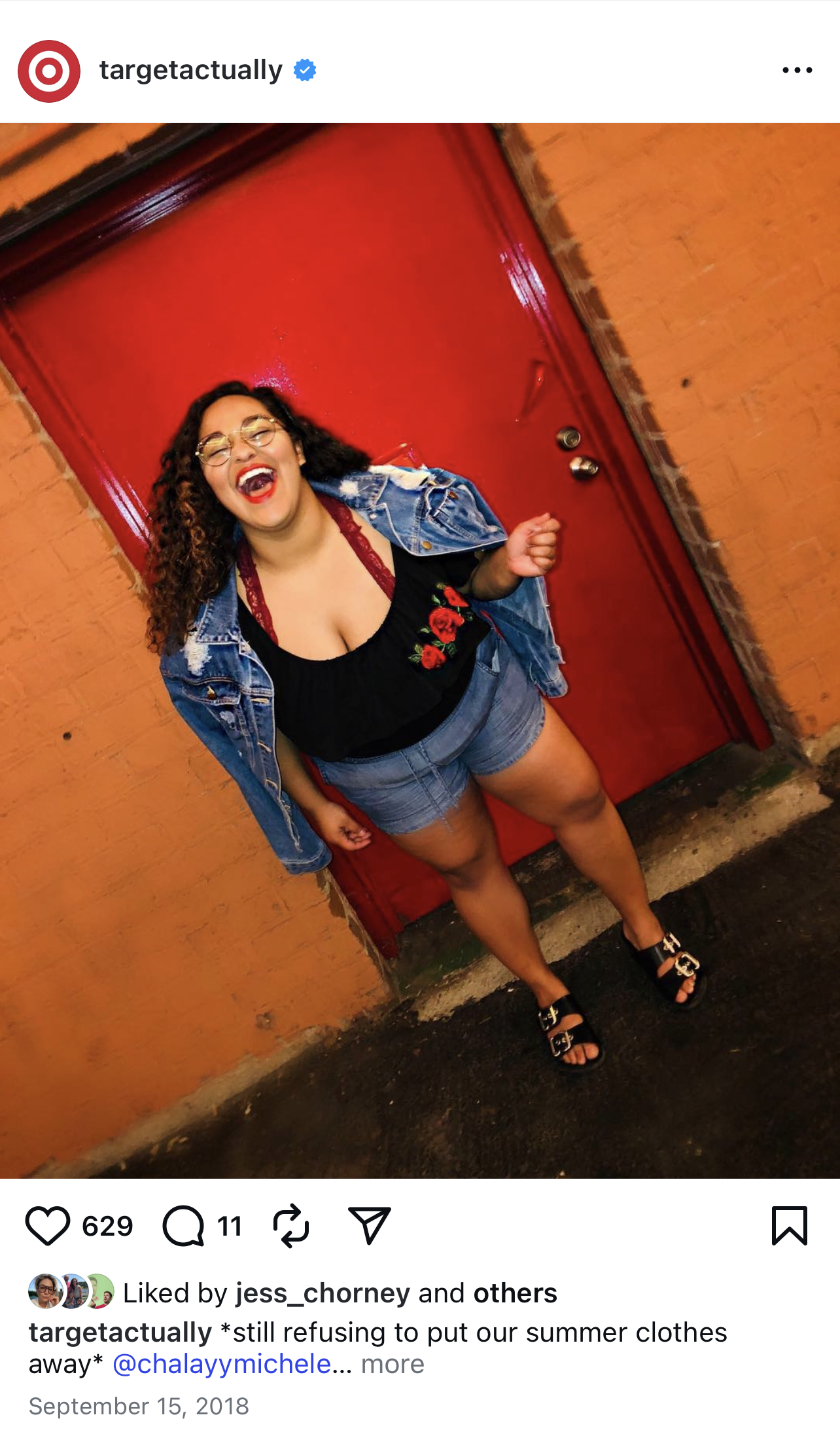 A woman with curly hair, glasses, and red lipstick, standing in front of a red door with a brick wall on one side. She is wearing a denim jacket, a black top with red floral embroidery, shorts, and black sandals. She is laughing with her mouth open.