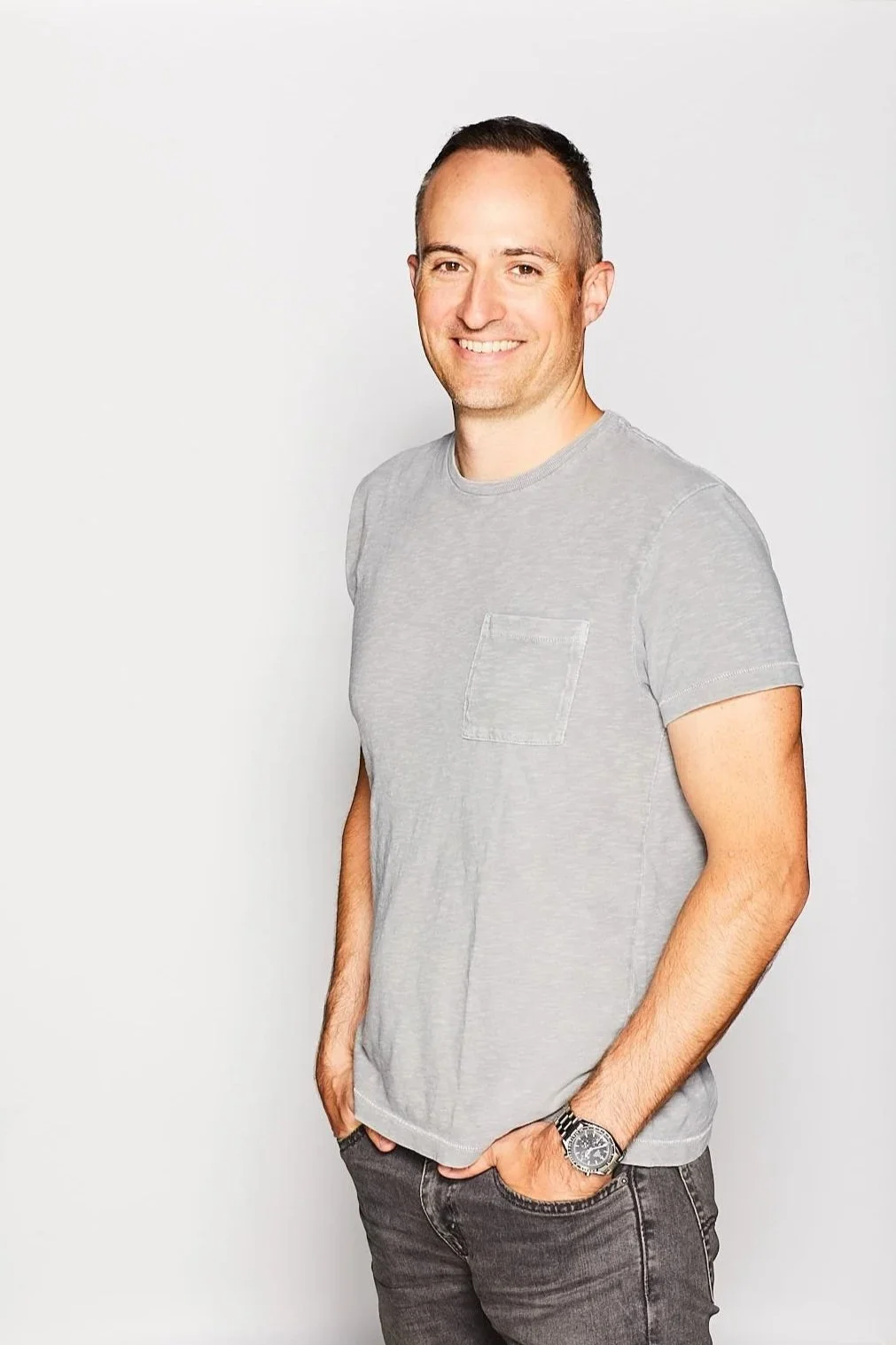 A smiling man with short dark hair wearing a light gray T-shirt with a pocket, dark jeans, and a wristwatch, standing against a plain white background.